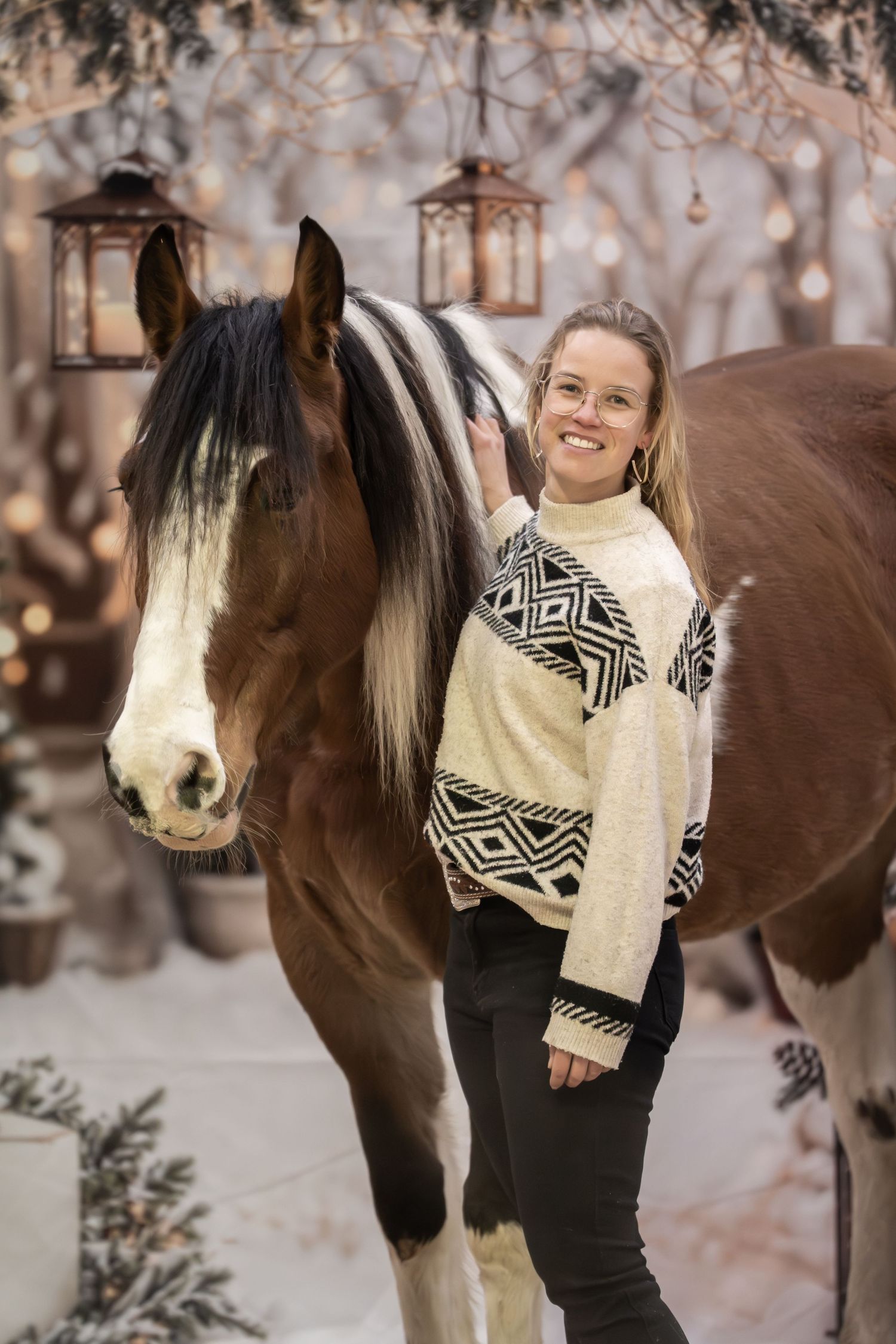 A winter scene with a brown and white horse standing beside someone in a black and white patterned sweater in the snow.