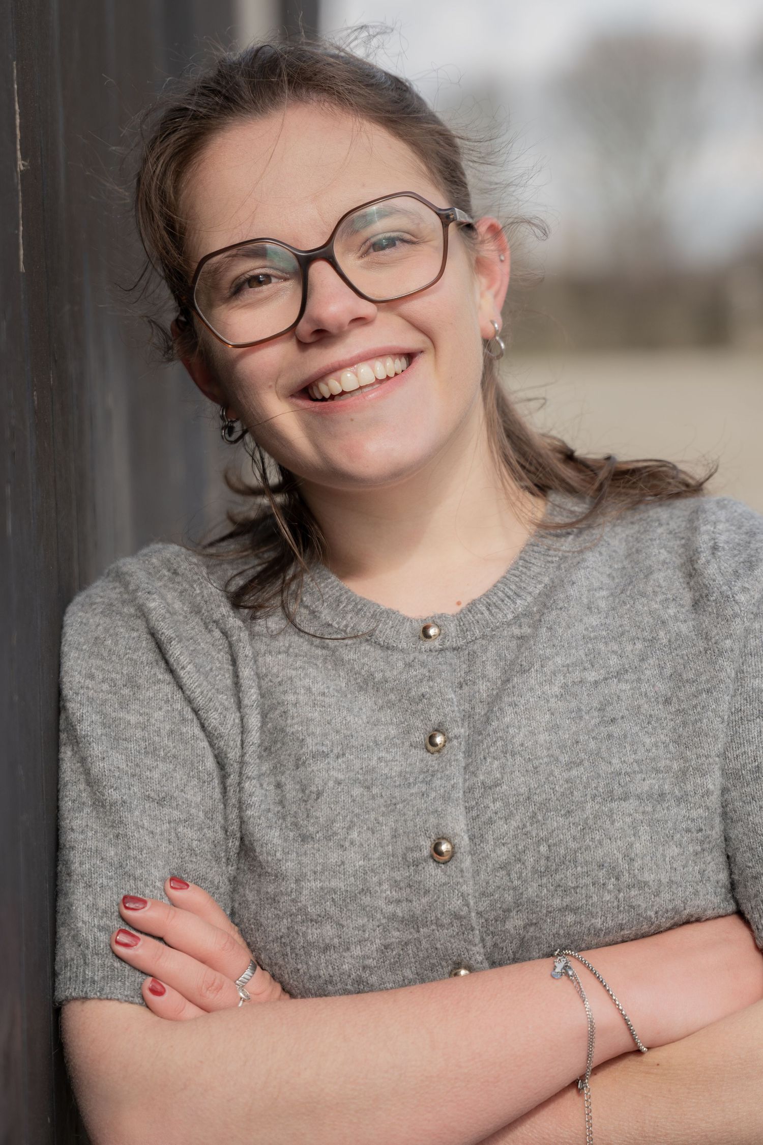 Smiling person in glasses wearing a gray cardigan sweater leaning against wall.