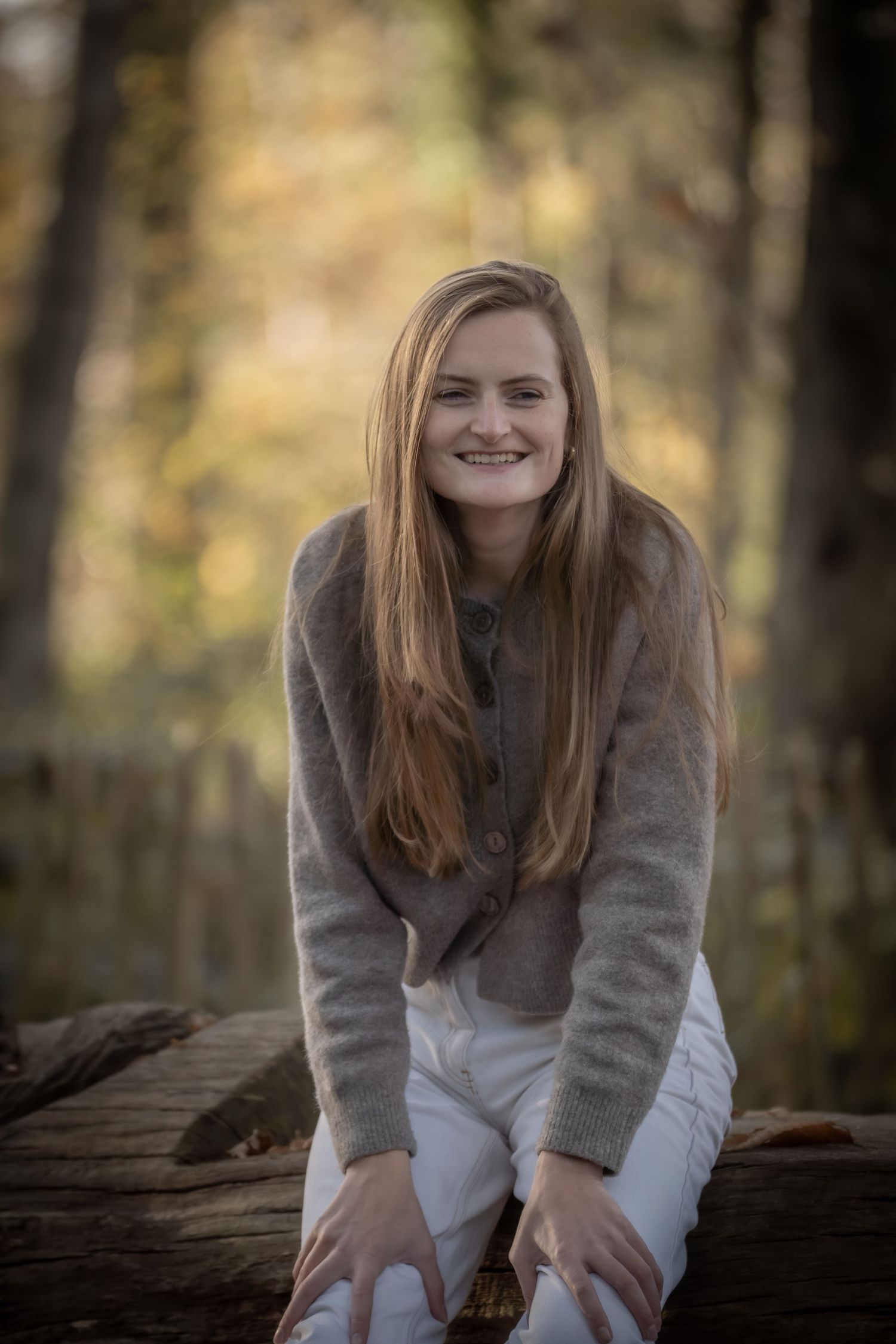 A person in a gray sweater and white pants sits on a fallen log in an autumn forest with blurred background.