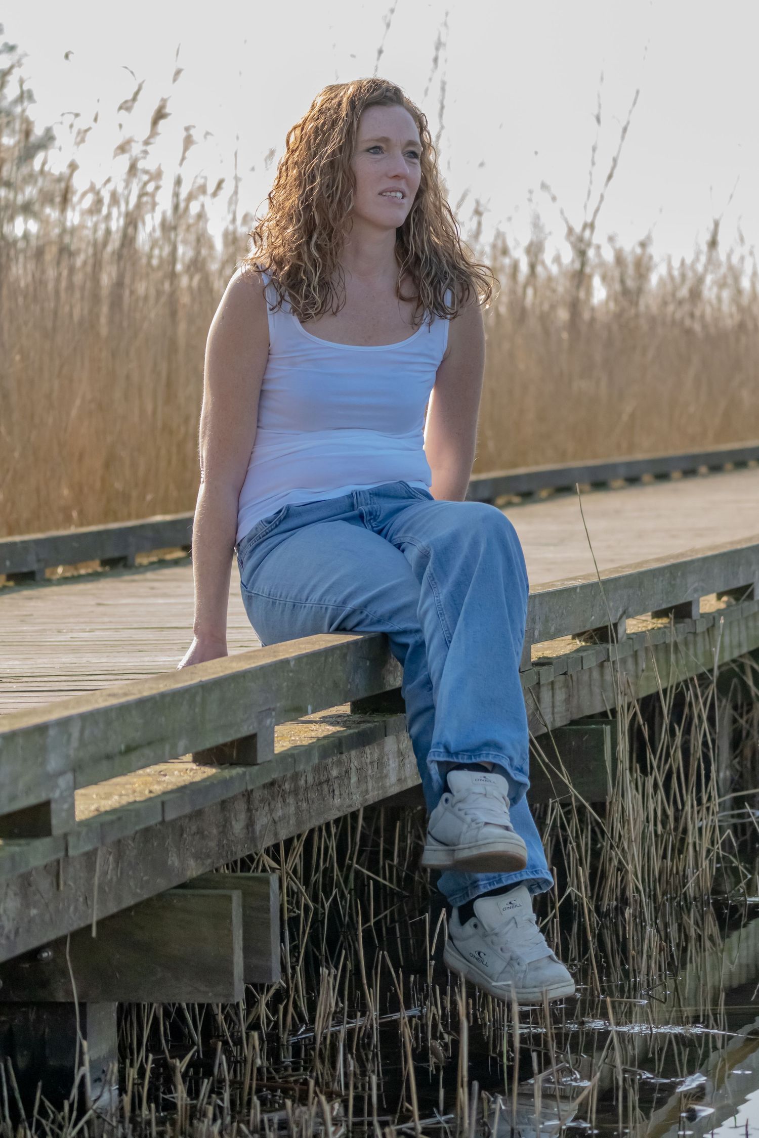 Person in casual attire sits relaxed on wooden dock with tall grass in background.