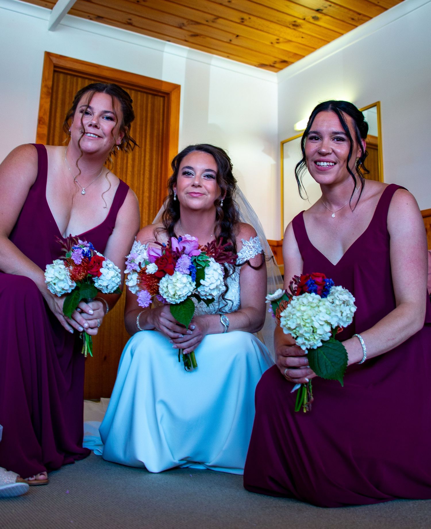 Three women in purple dresses holding floral bouquets in indoor setting.