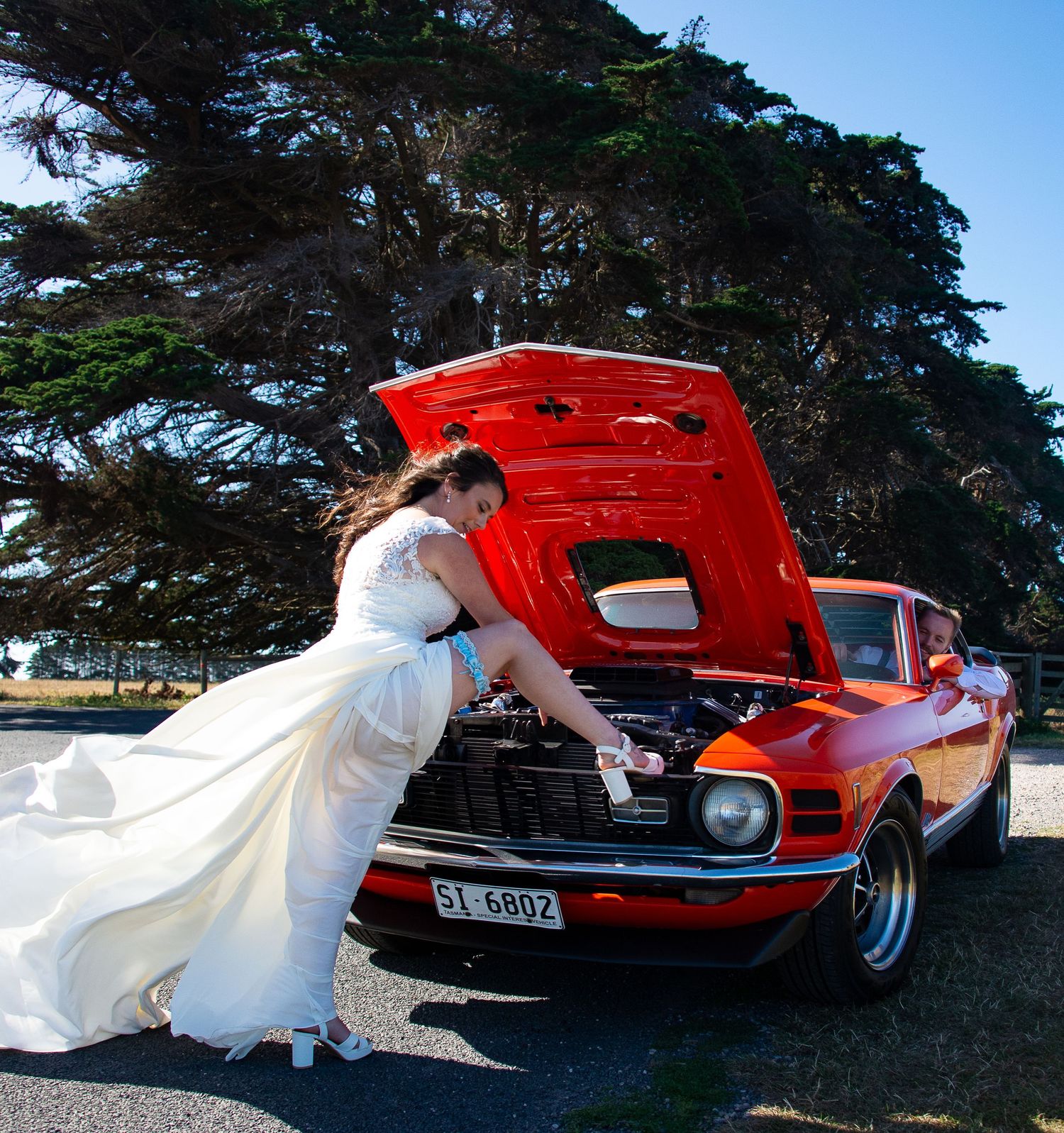 Bride in white dress checking under hood of classic red Mustang car.