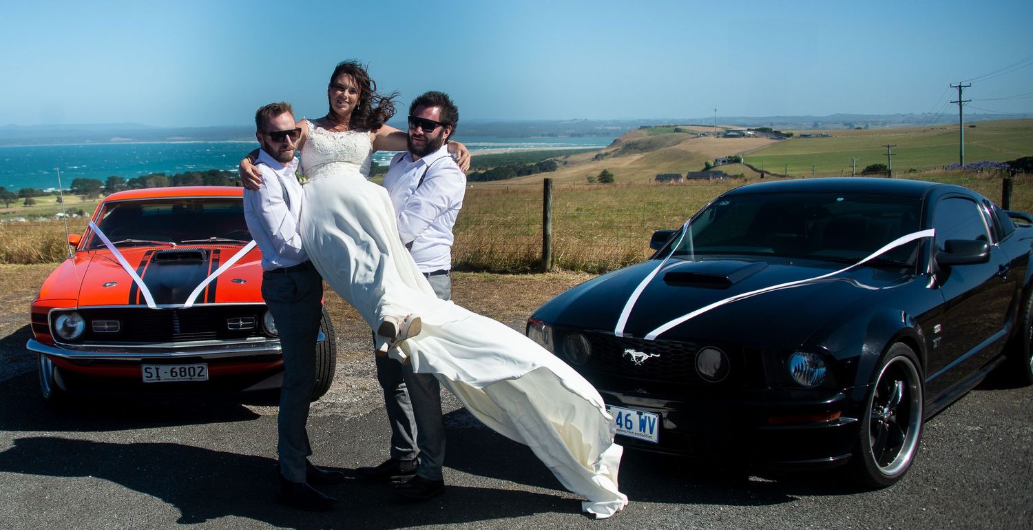 Wedding party posing with classic cars, bride's dress flowing in the wind.