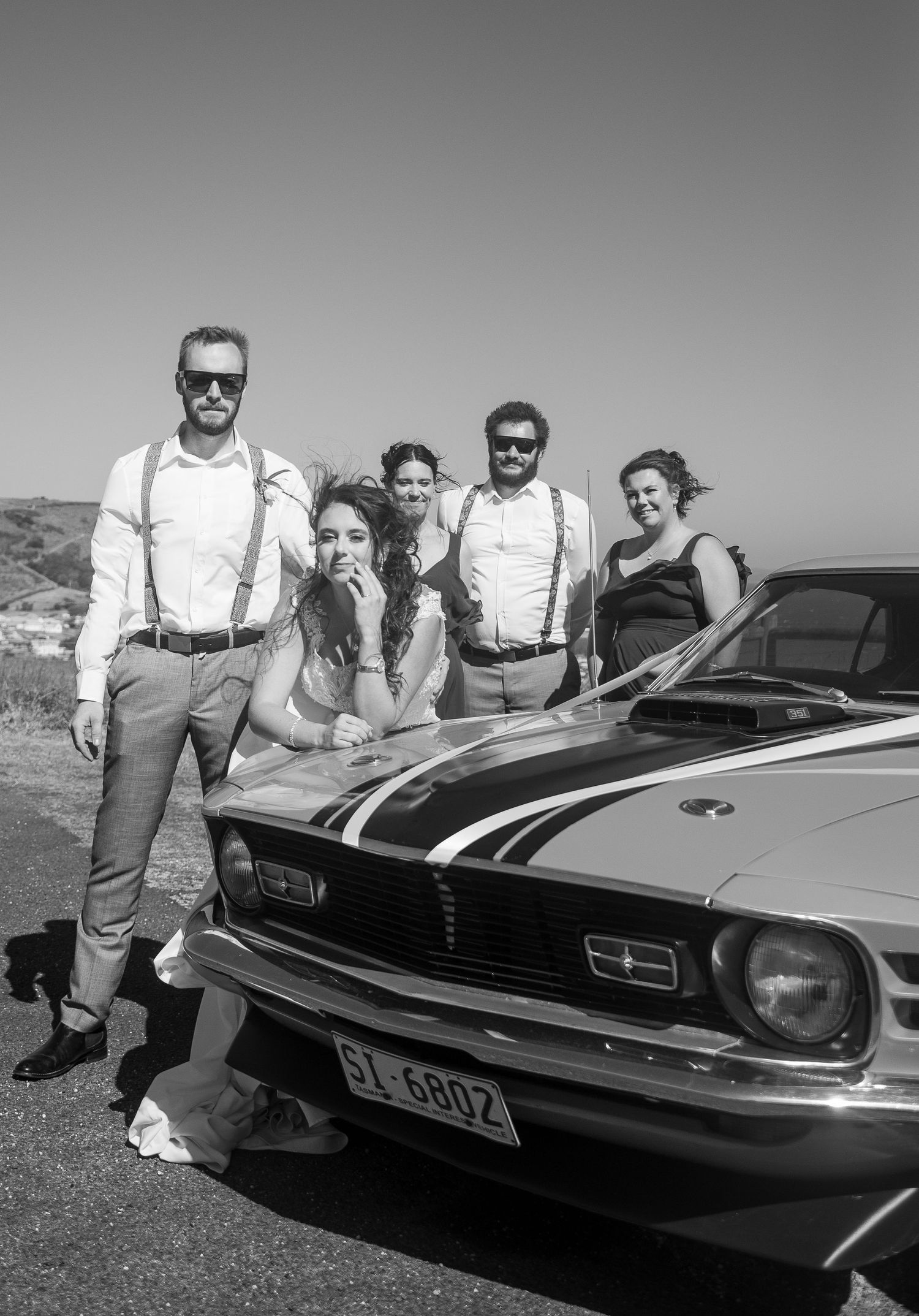 Wedding party in formal attire posing with classic Mustang car in black and white photo.