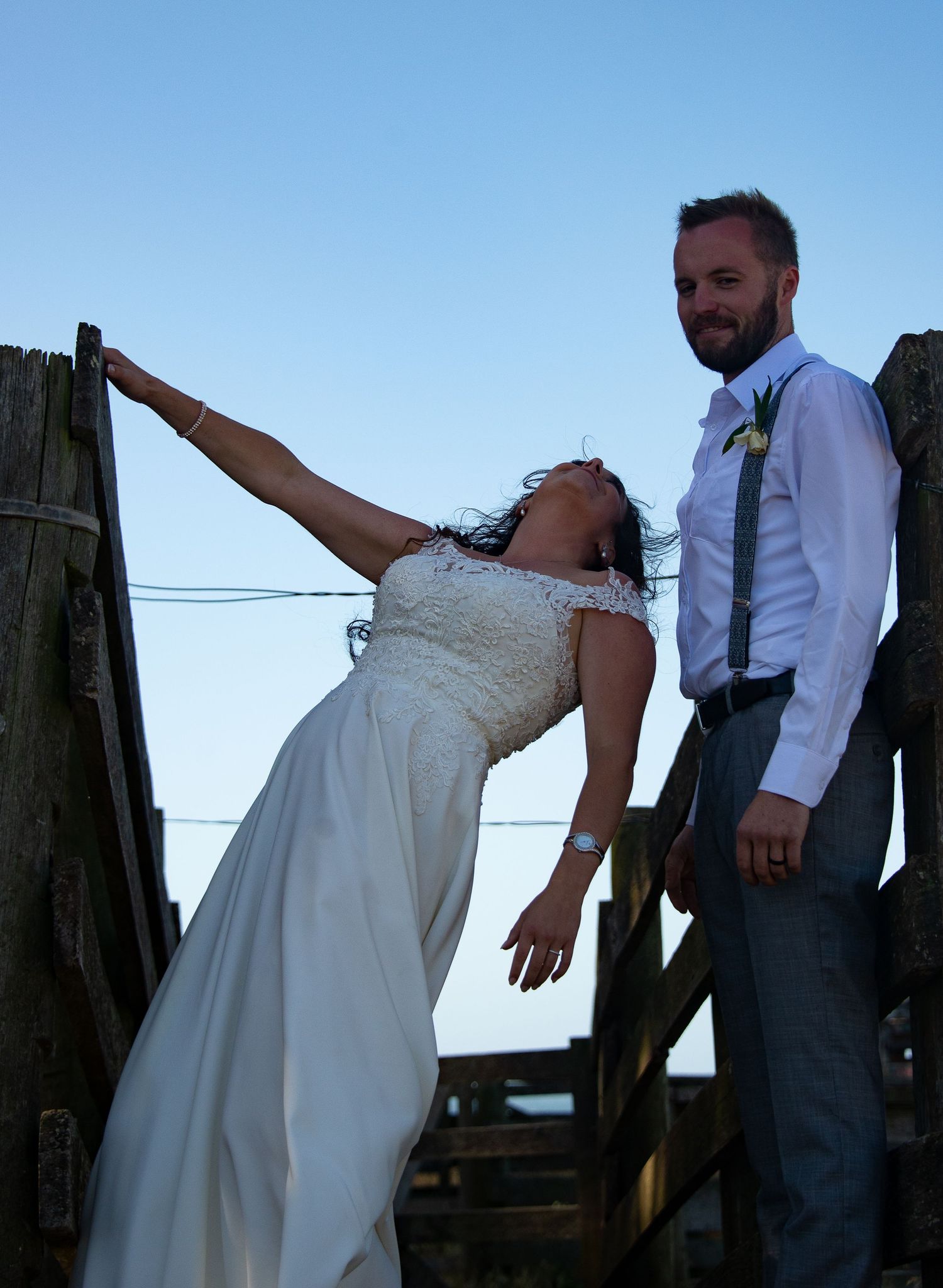 Artistic wedding pose with bride leaning back on wooden deck railing at dusk.