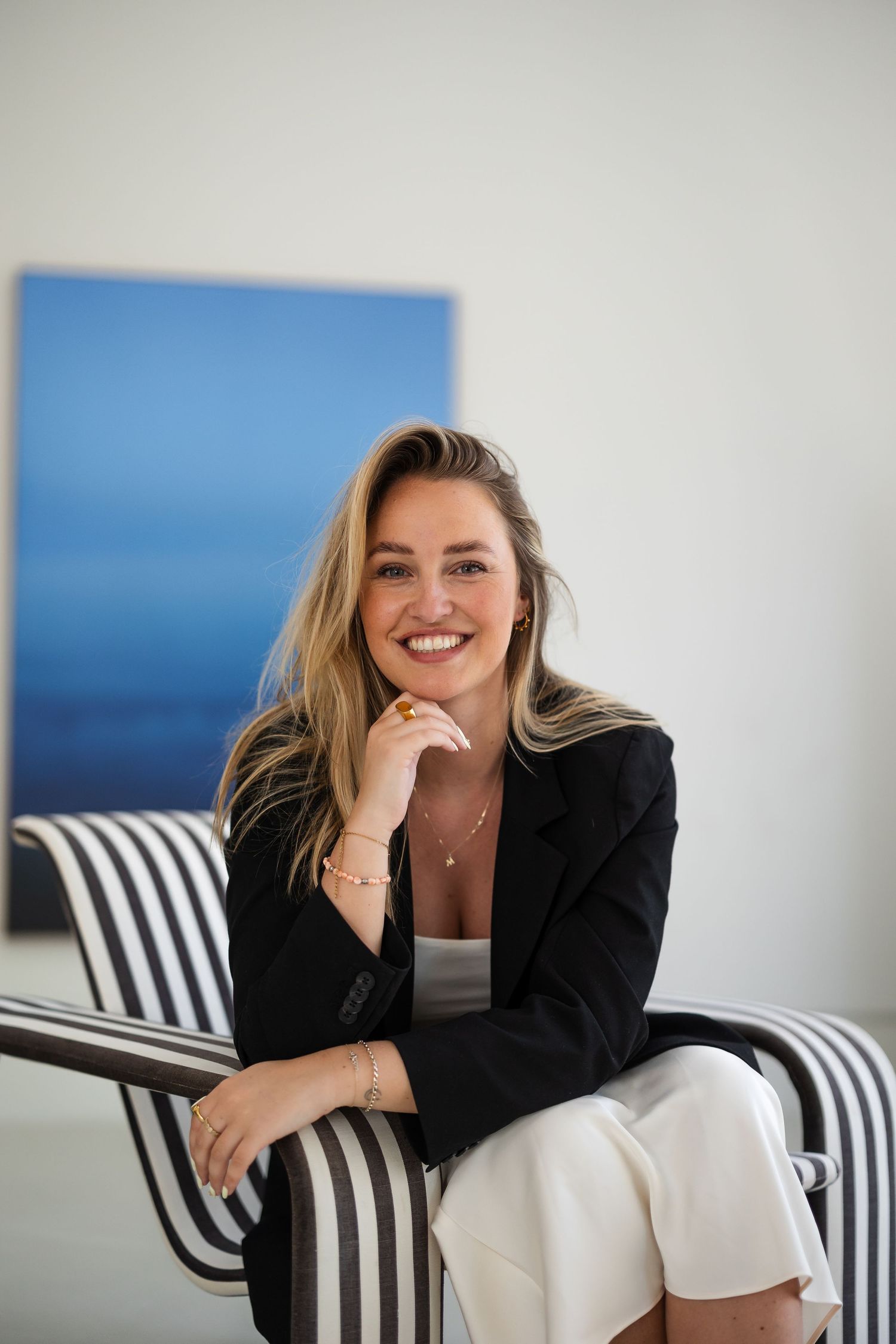 Professional portrait of someone wearing a black blazer and white pants seated on a striped chair against a coastal backdrop.