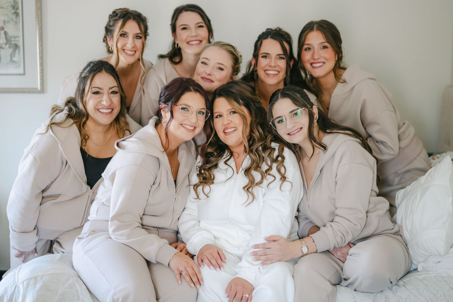 Bride in white robe smiling with eight bridesmaids in matching beige loungewear, posing together on a bed. - Youngblood Vineyard Wedding