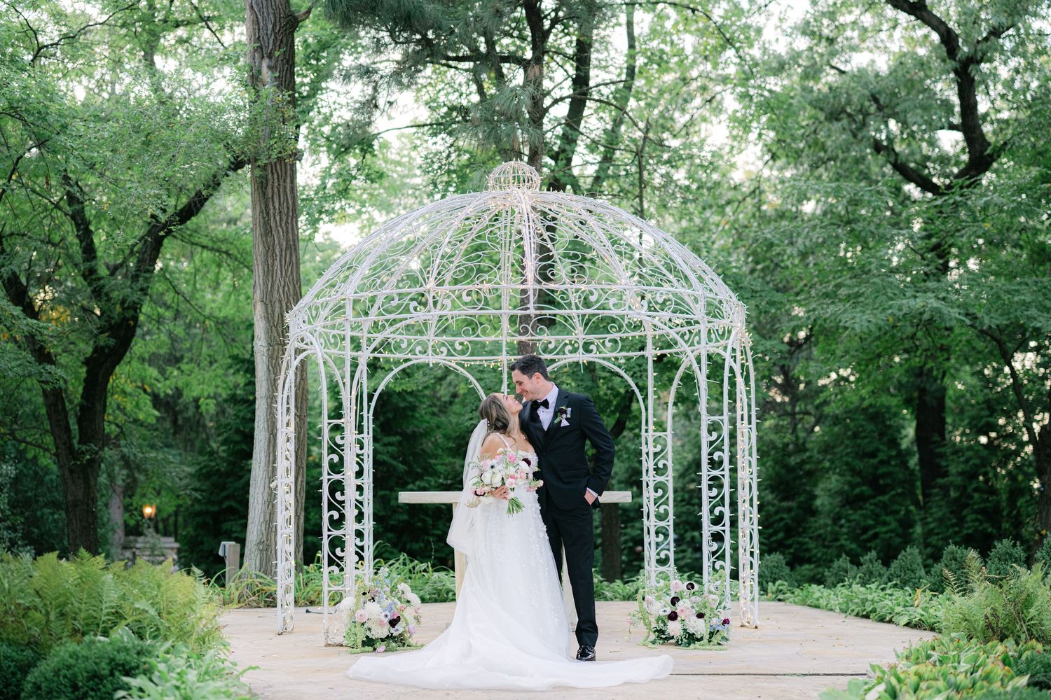A wedding ceremony takes place under a white metal gazebo surrounded by lush green trees in a garden setting. - Meyer's Castle Events