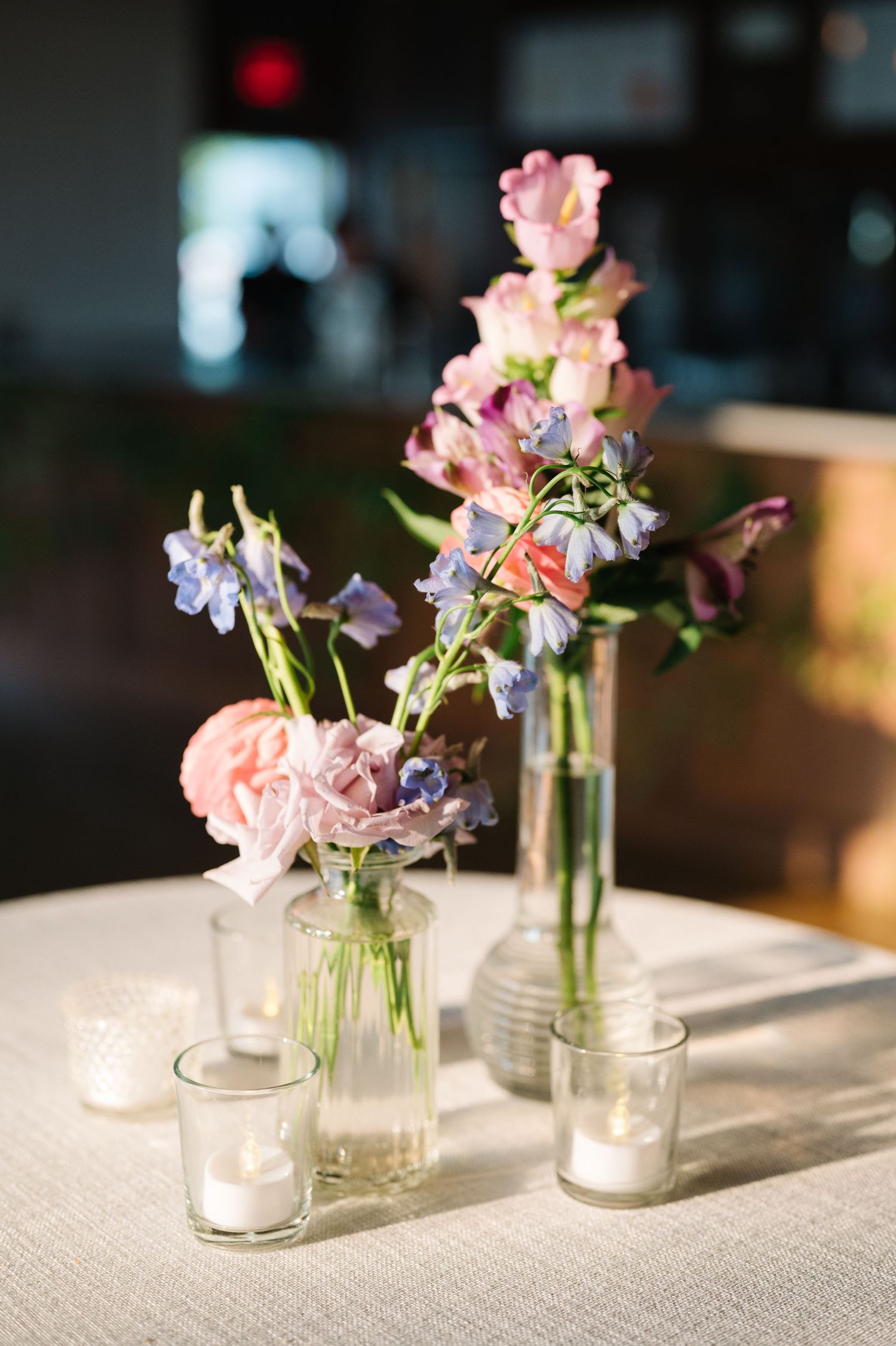 Simple floral centerpiece with pink carnations and purple flowers in glass bottles alongside tea light candles on light fabric.