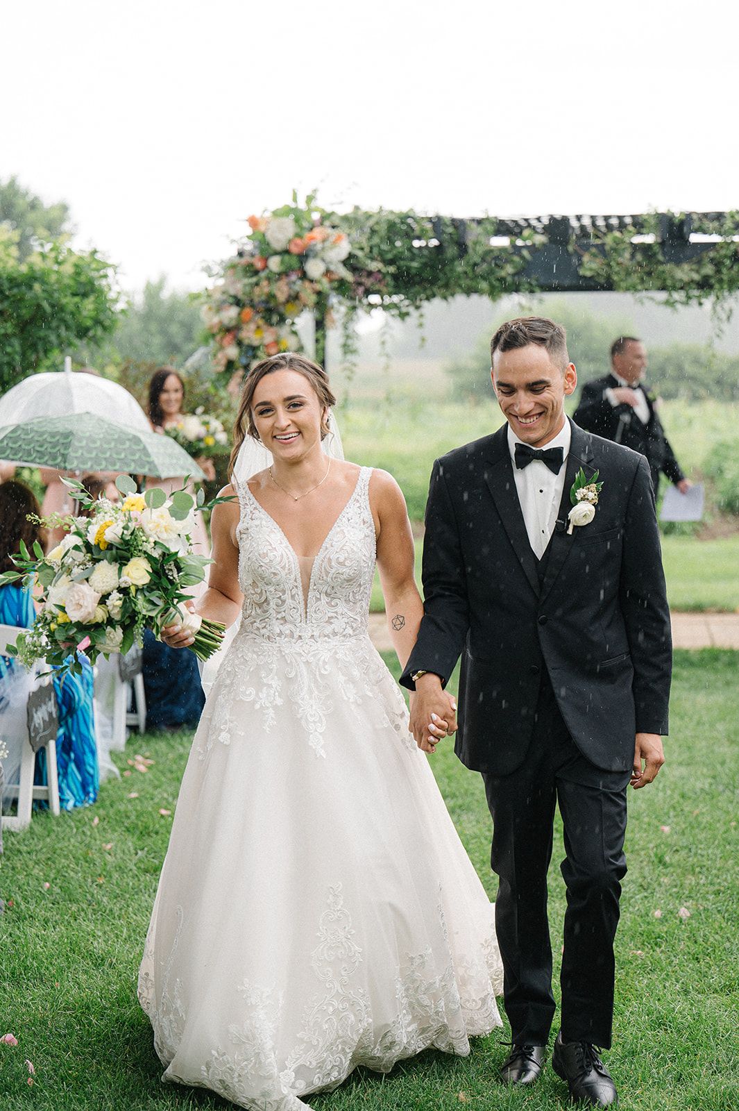 Bride and groom walk hand in hand after outdoor wedding ceremony, bride holding floral bouquet. - Heritage Prairie Farm Wedding