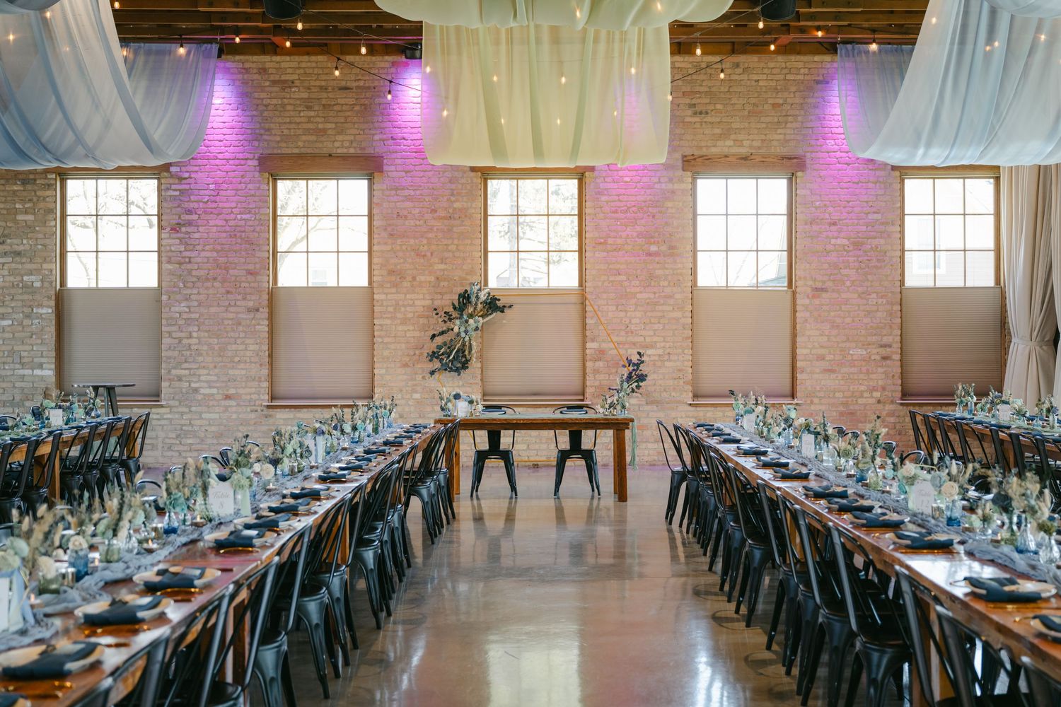 Long banquet tables with blue chairs are set up in an industrial-style venue with exposed brick and draped fabric. - The Brix on the Fox