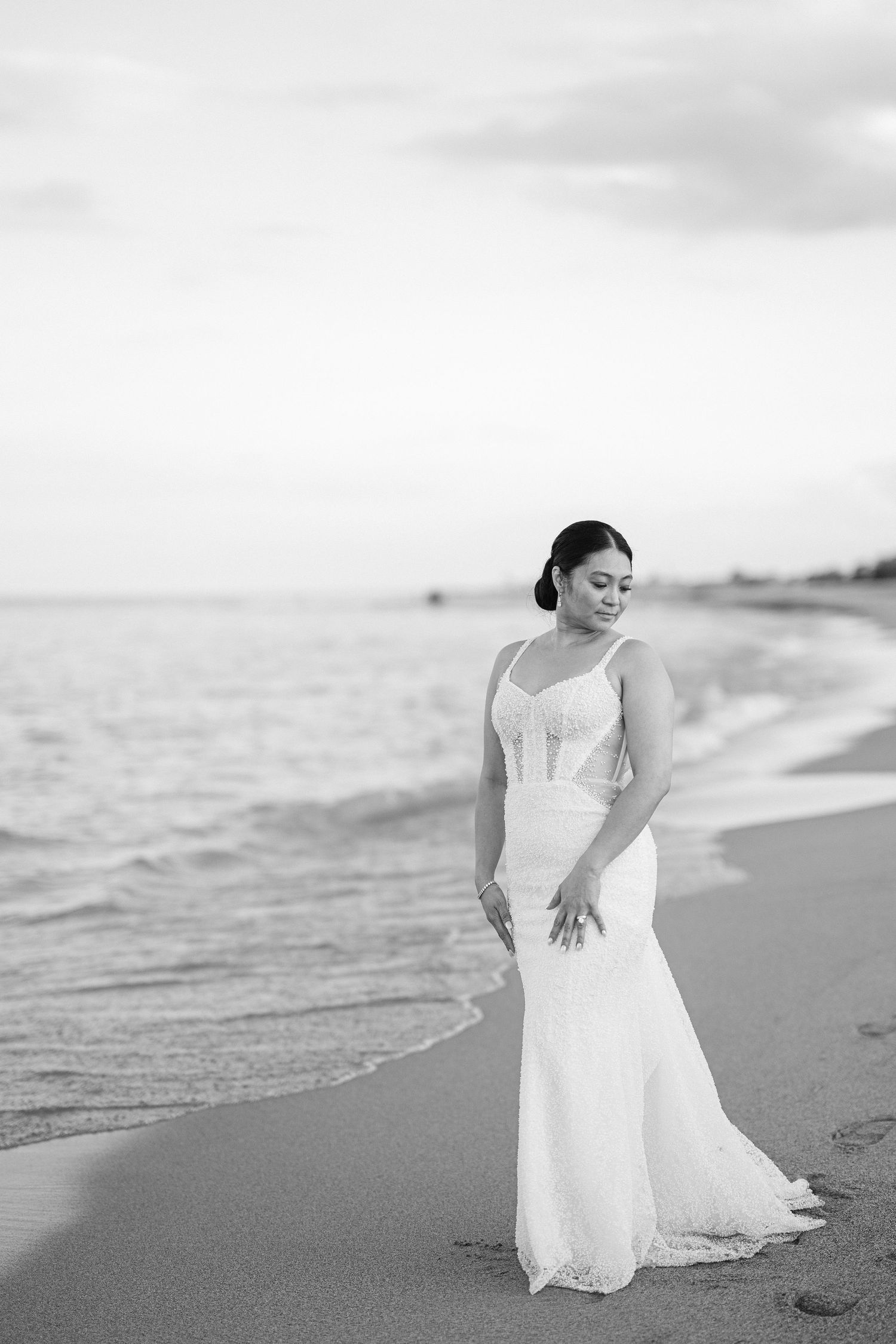A bride in a fitted white gown poses alone on a beach at sunset.