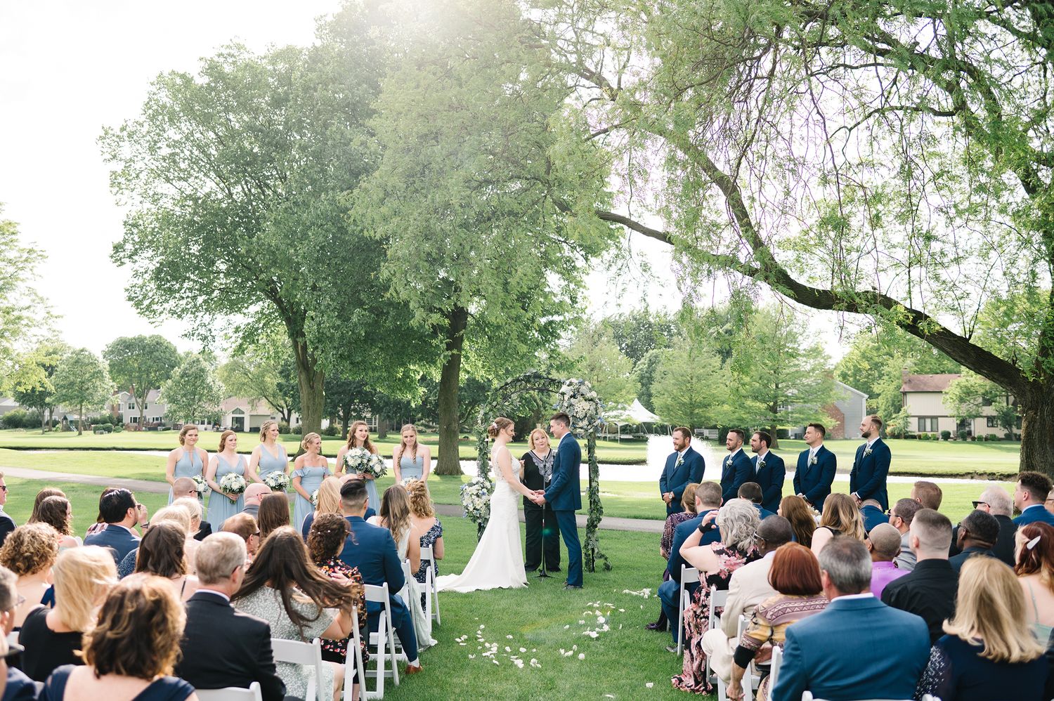 An outdoor wedding ceremony takes place on a lush green lawn under towering trees with guests seated in white chairs.