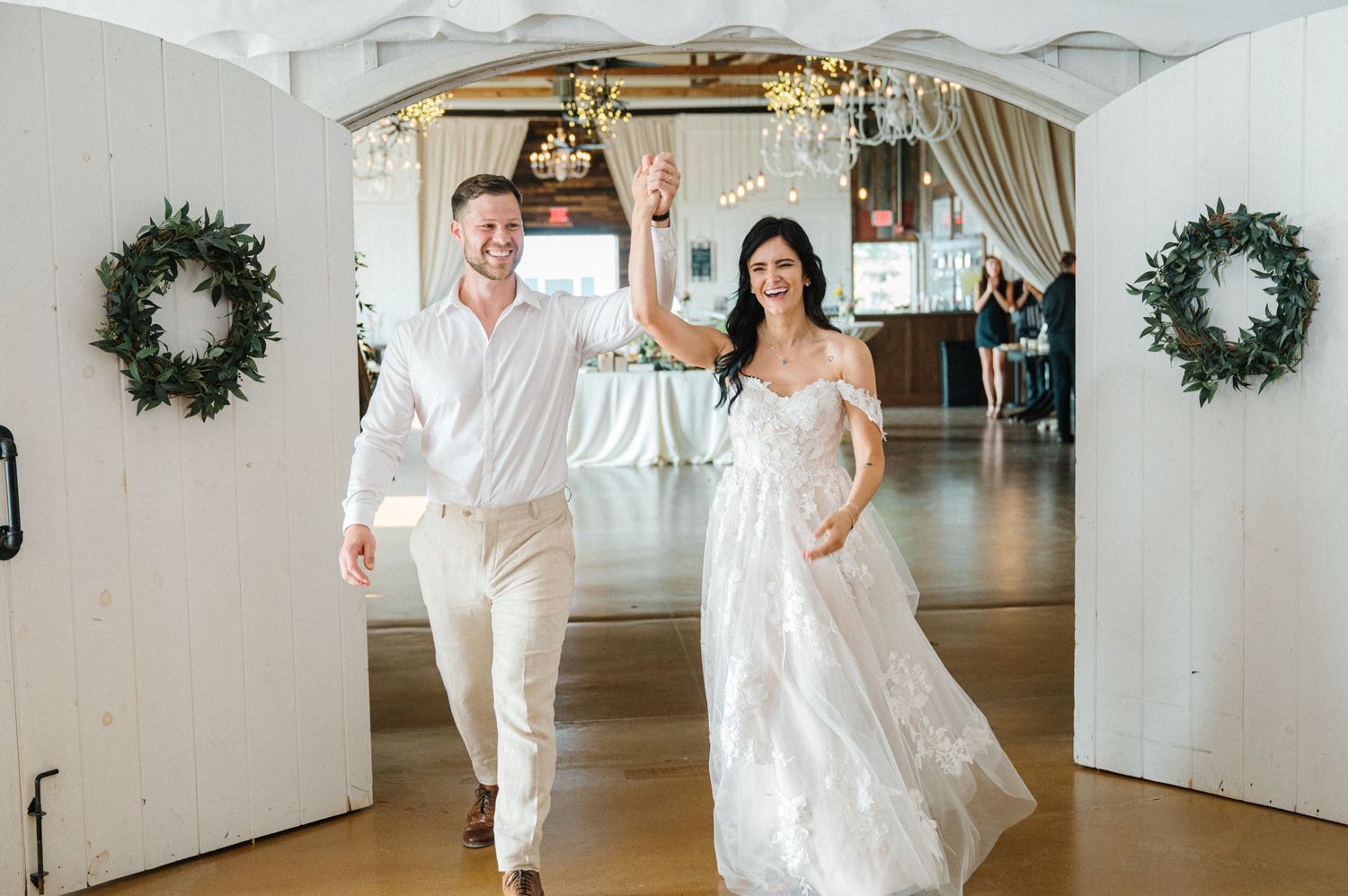 Happy newlyweds in white attire joyfully dancing and celebrating in an elegant venue decorated with green wreaths.