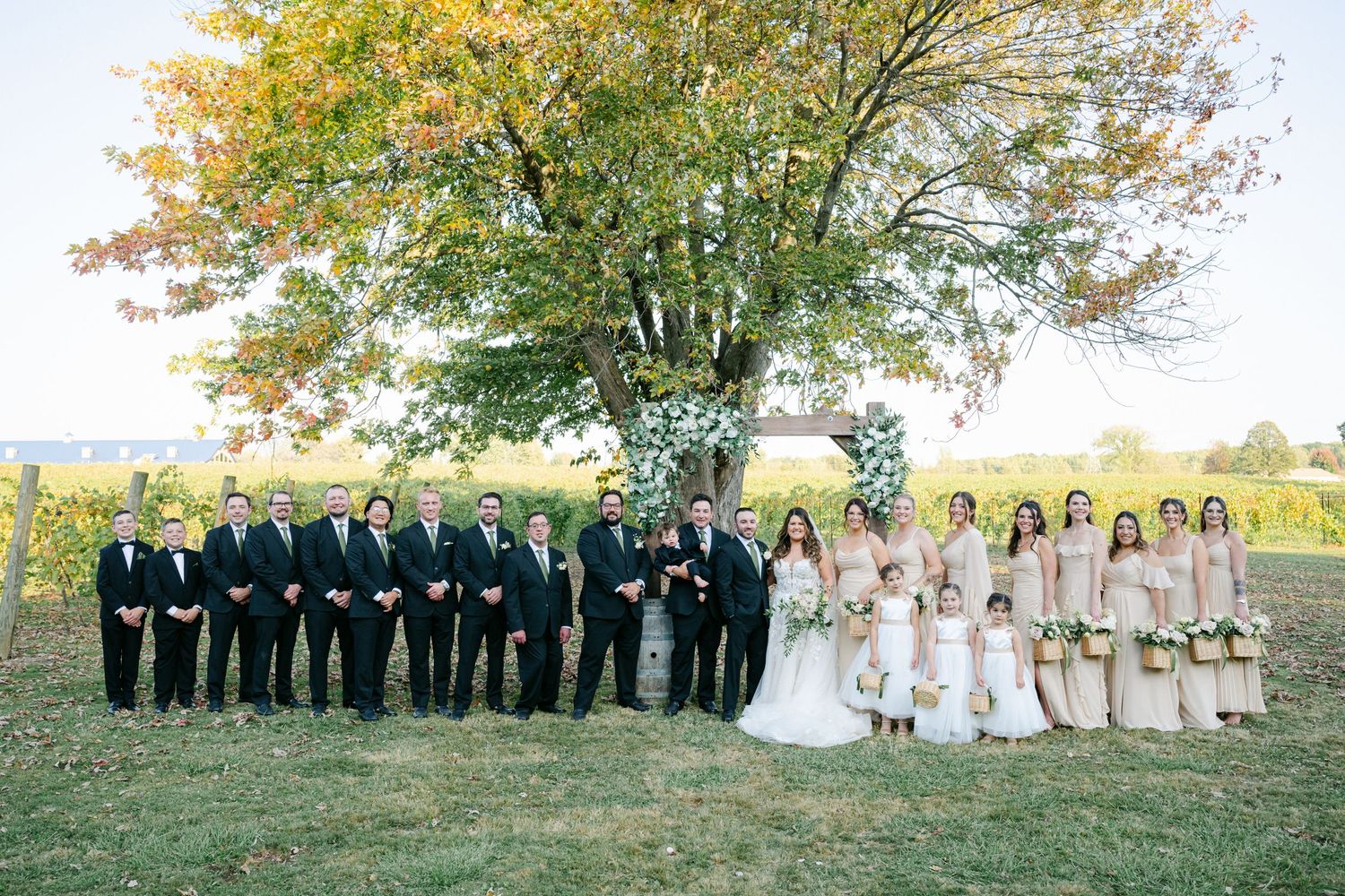 Wedding party poses outdoors near a floral arch and large tree, with groomsmen and bridesmaids. - Youngblood Vineyard Wedding