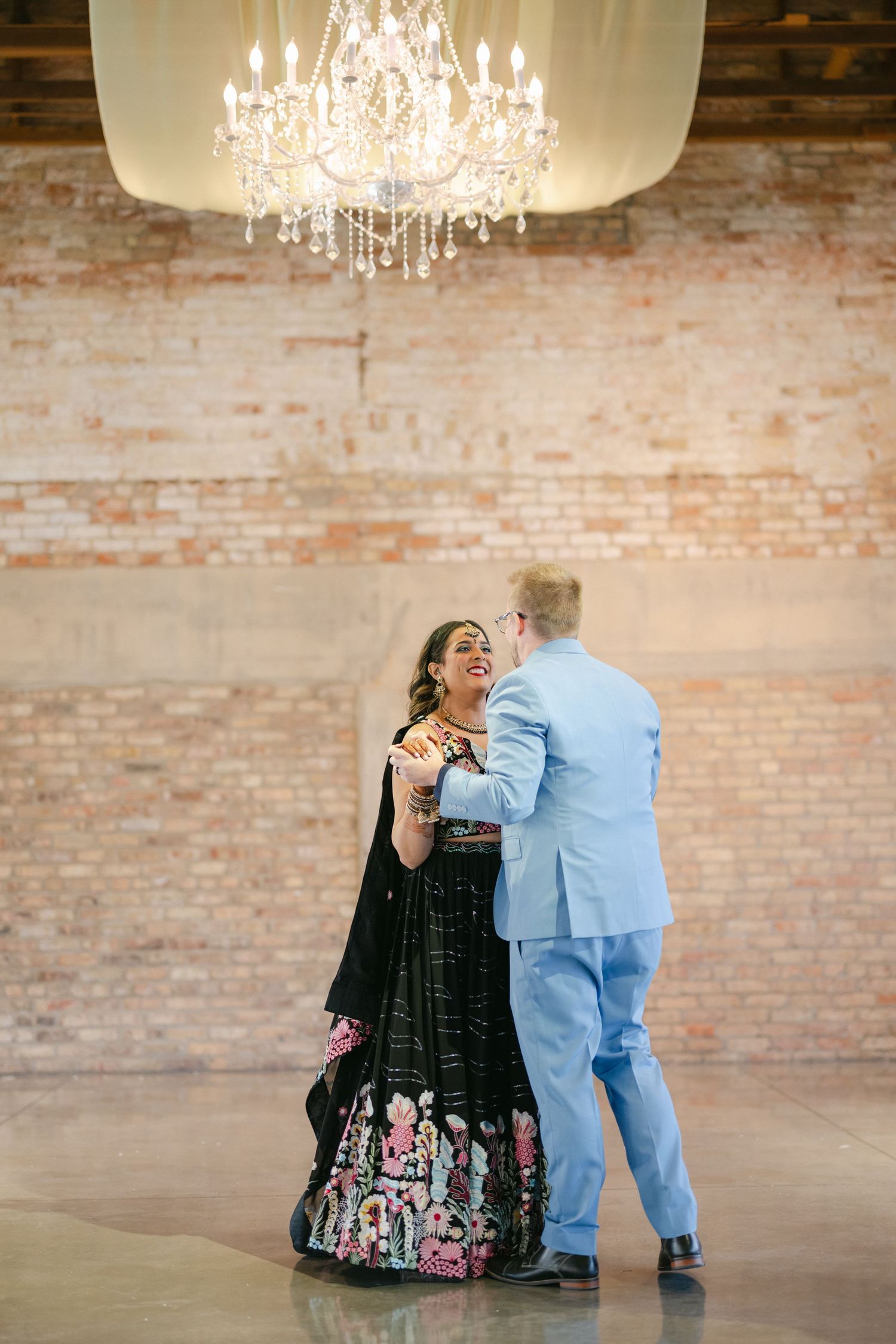 A couple dances beneath an elegant crystal chandelier in a rustic brick-walled venue. - The Brix on the Fox