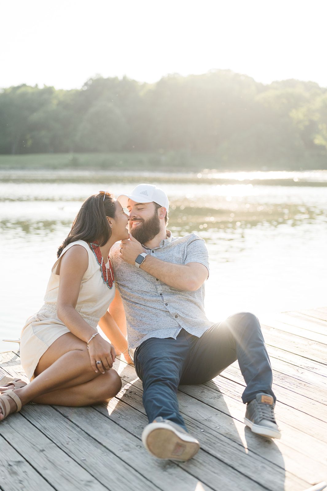 Couple sitting on a wooden dock by a lake, sharing a romantic moment in golden sunset light. - Wheaton Engagement Session