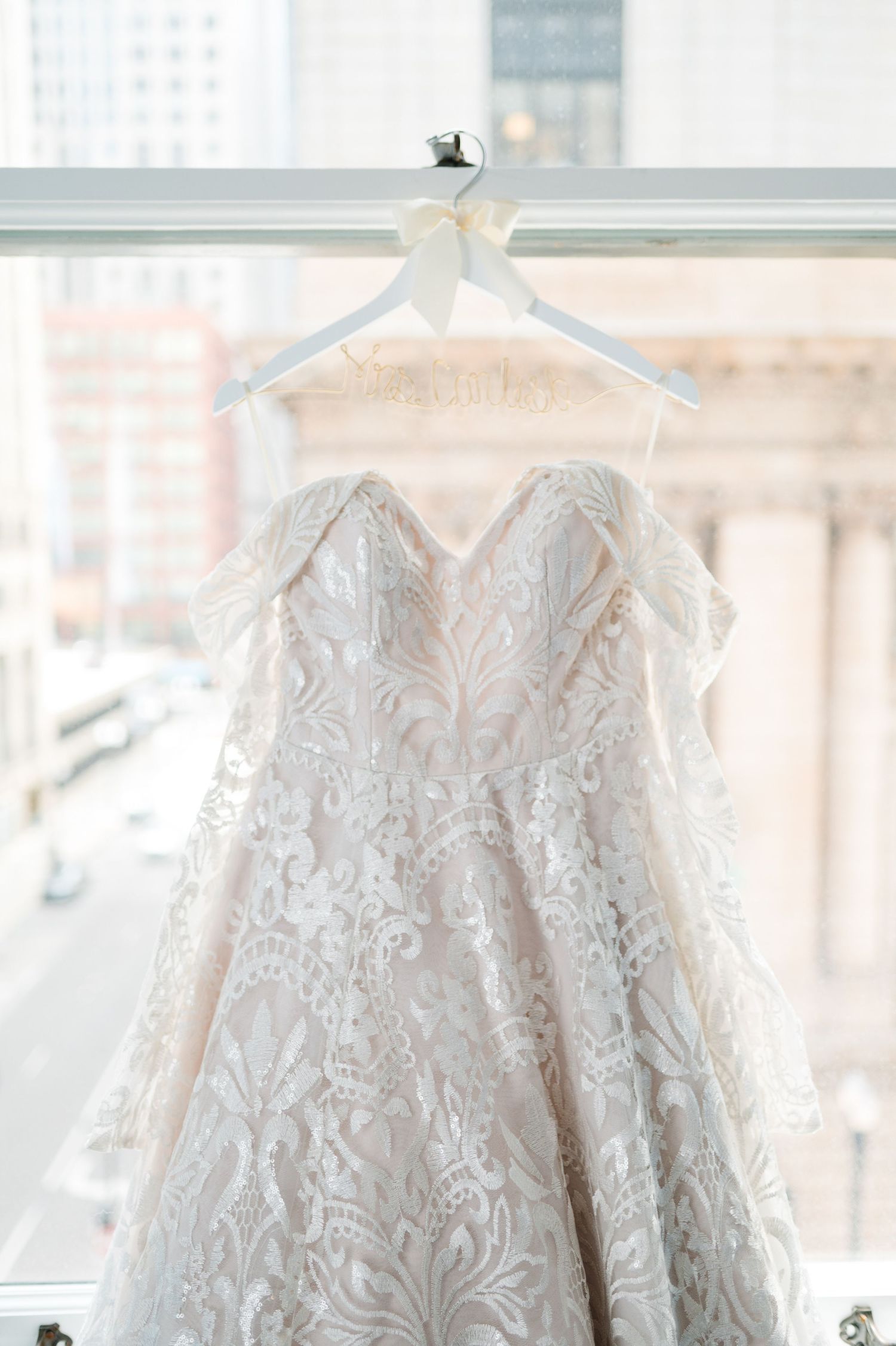 A delicate white lace wedding dress with intricate floral patterns hangs from a white hanger. - Chicago Wedding Photographer