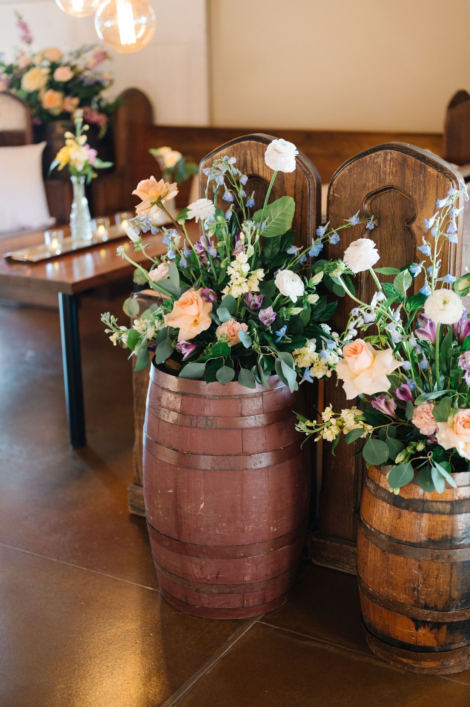 Rustic wooden barrels decorated with peach roses, white flowers and greenery arrangements in an indoor venue setting.
