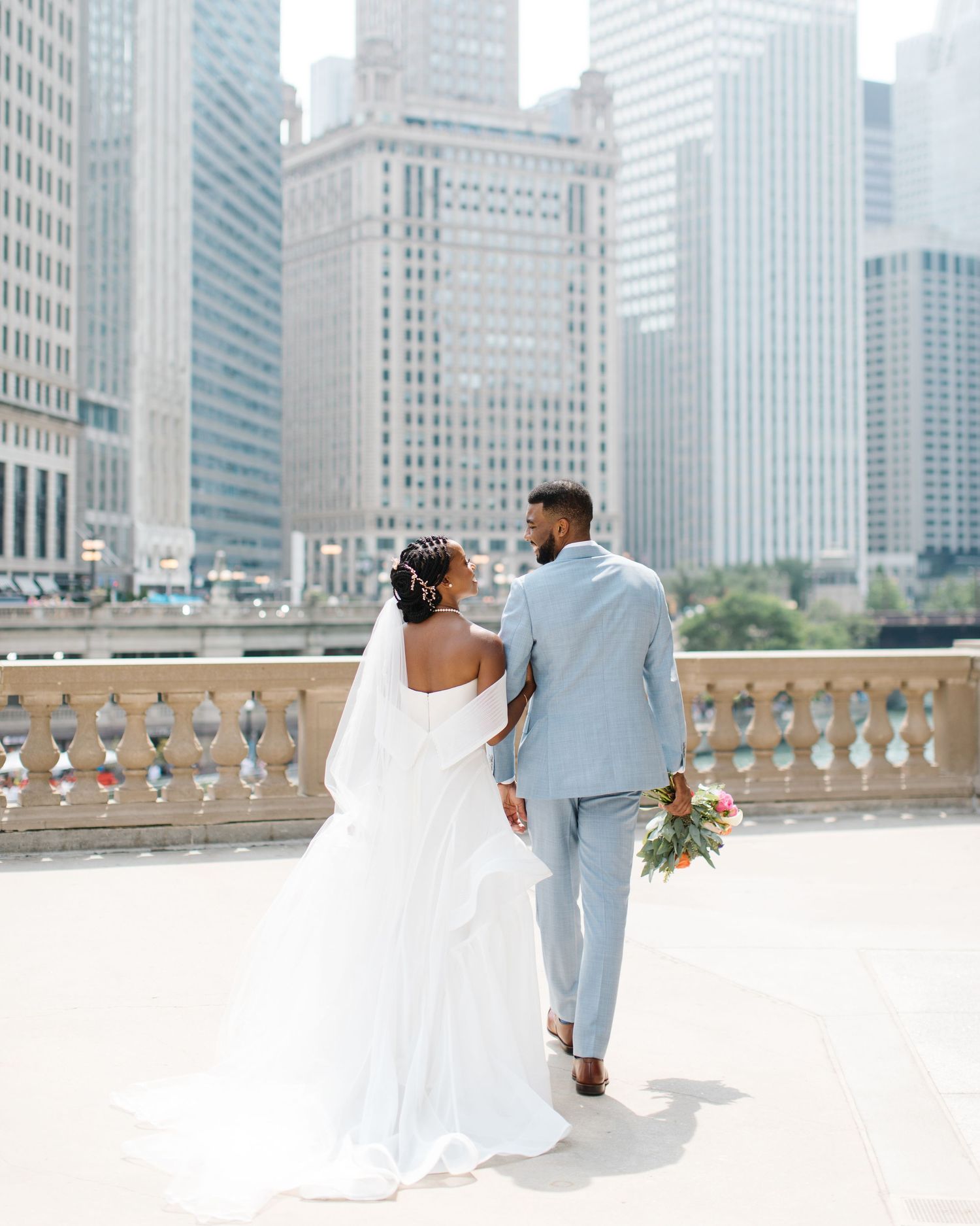 Wedding couple walking along stone balcony with Chicago skyscrapers in background.