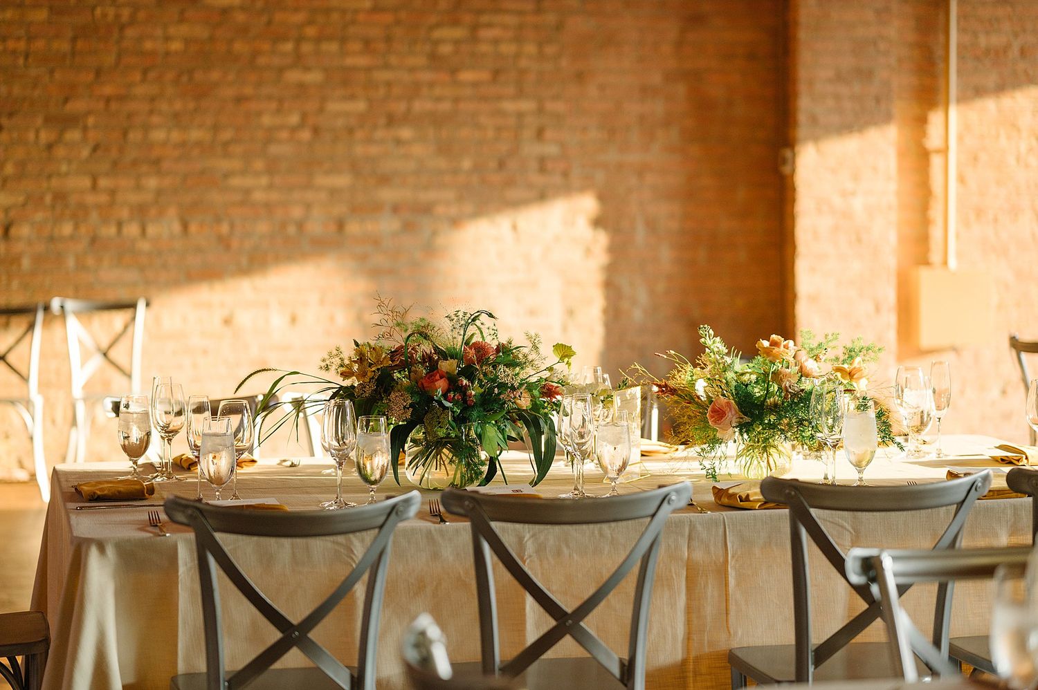Rustic wedding reception table with greenery centerpieces and cross-back chairs against exposed brick wall in warm sunlight.