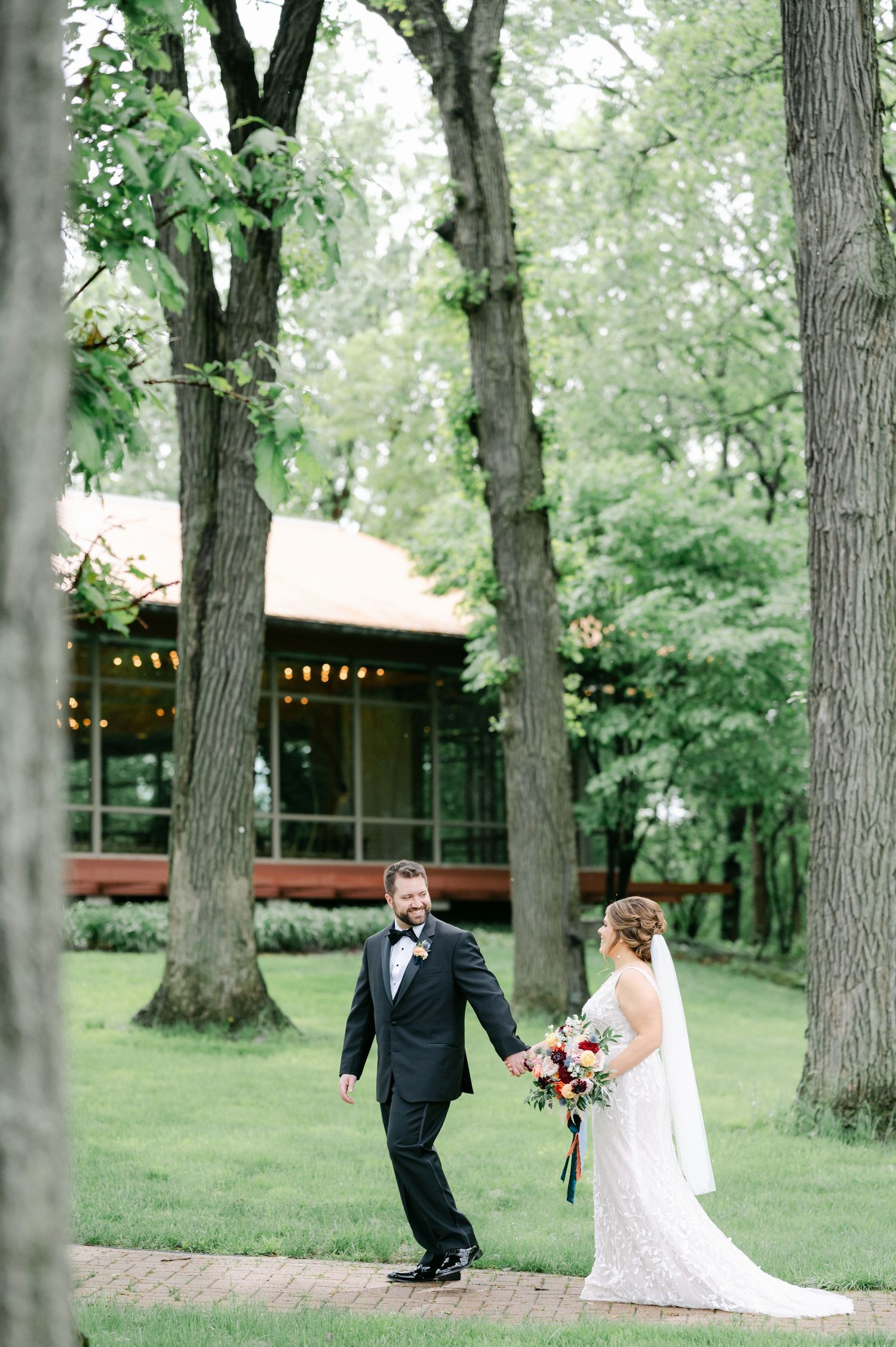 A newlywed couple walks hand in hand through a wooded outdoor venue with string lights and a rustic background. - Hyatt Lodge Oak Brook