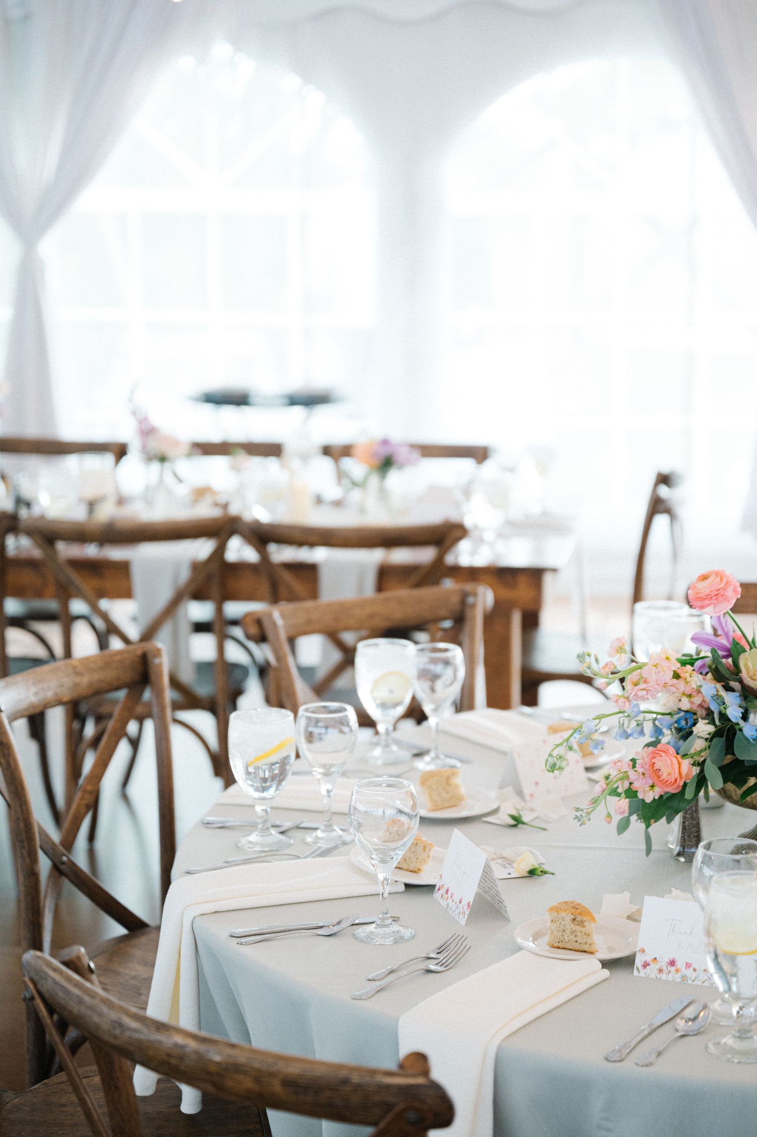 An elegant wedding reception table setting with wooden chairs, white linens, and soft pink floral centerpieces under a draped tent.