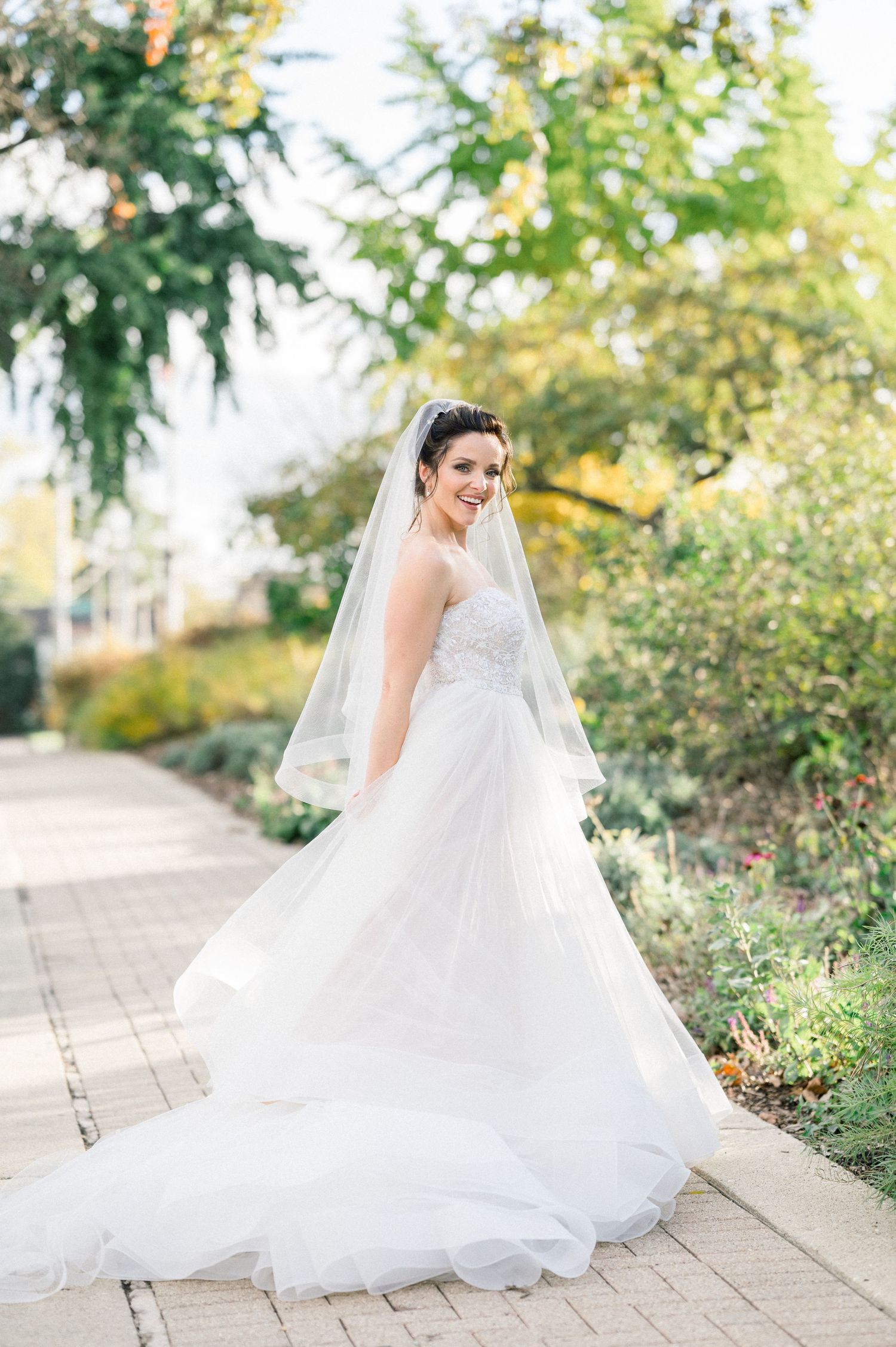 Bride in elegant white gown with cathedral veil turns on stone pathway.