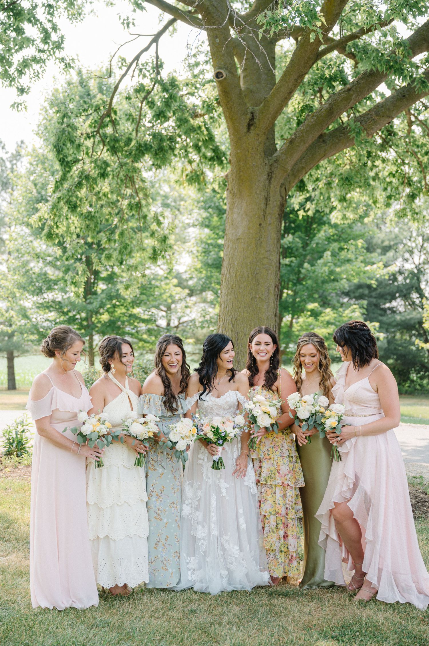 Group in pastel and floral dresses posing under large tree with bouquets. - Venue 5126 Wedding