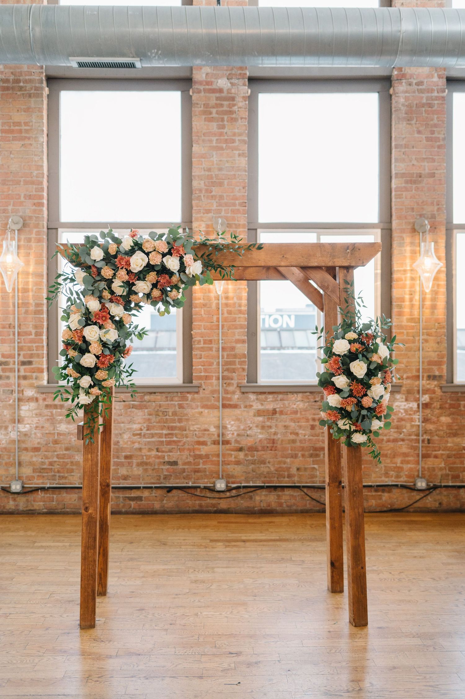 Wooden posts adorned with lush peach and white floral arrangements line a rustic brick-walled venue. - Chicago's Western Suburbs Wedding