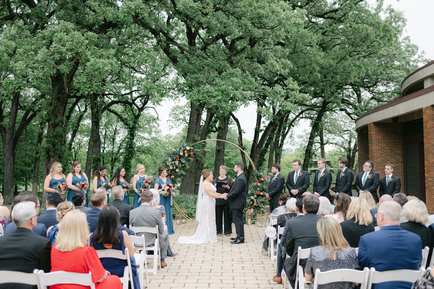 Outdoor wedding ceremony guests seated in white chairs under green trees with rustic wooden arch at front. - Hyatt Lodge Oak Brook