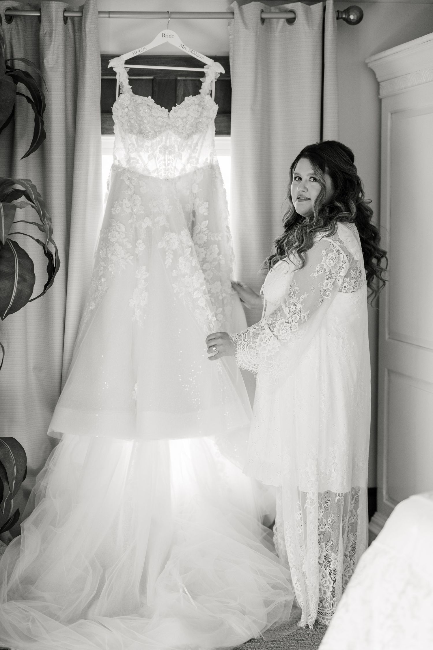Bride in lace robe admiring her elegant strapless ball gown wedding dress hanging by a window. - Youngblood Vineyard Wedding