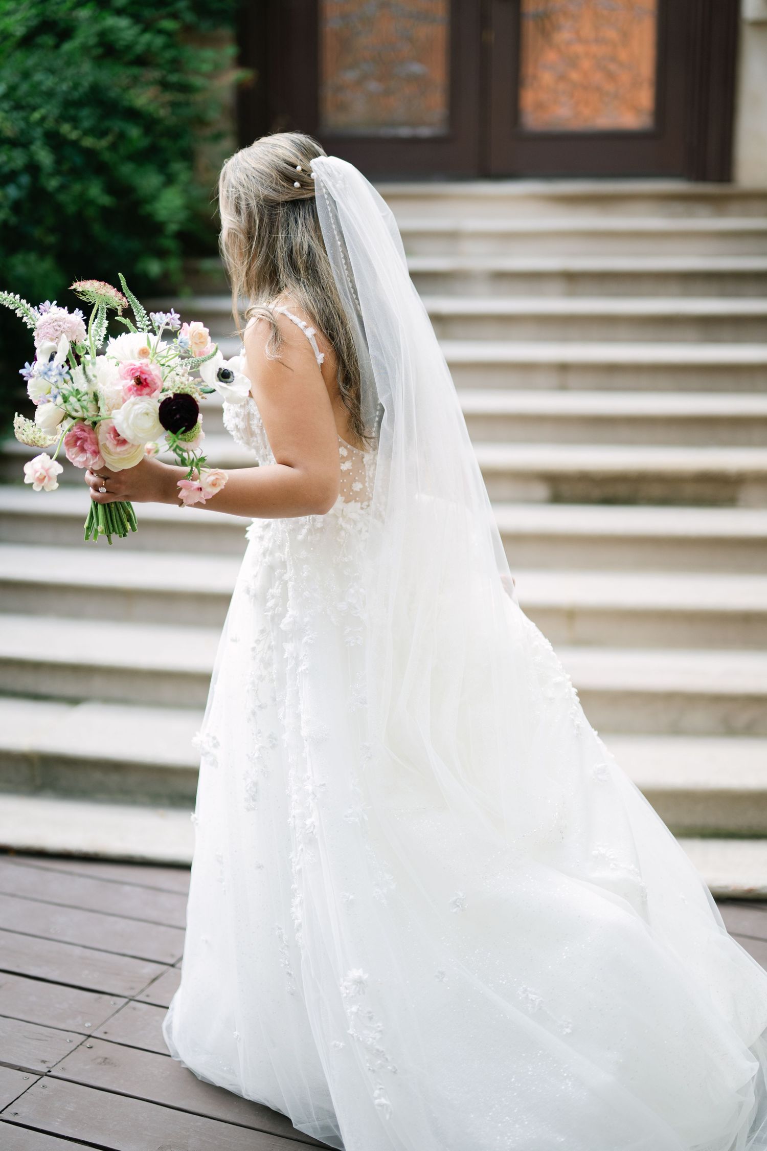 A bride in a flowing white wedding dress and veil holds a pink and white floral bouquet on stone steps. - Meyer's Castle Events