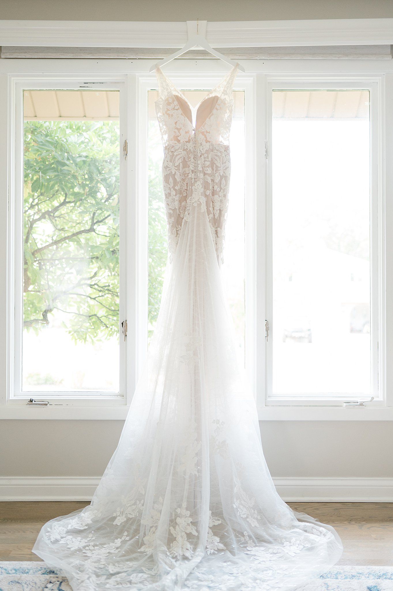 A delicate white lace wedding dress hangs in front of bright windows in a sunlit room. - The Bridge Lemont