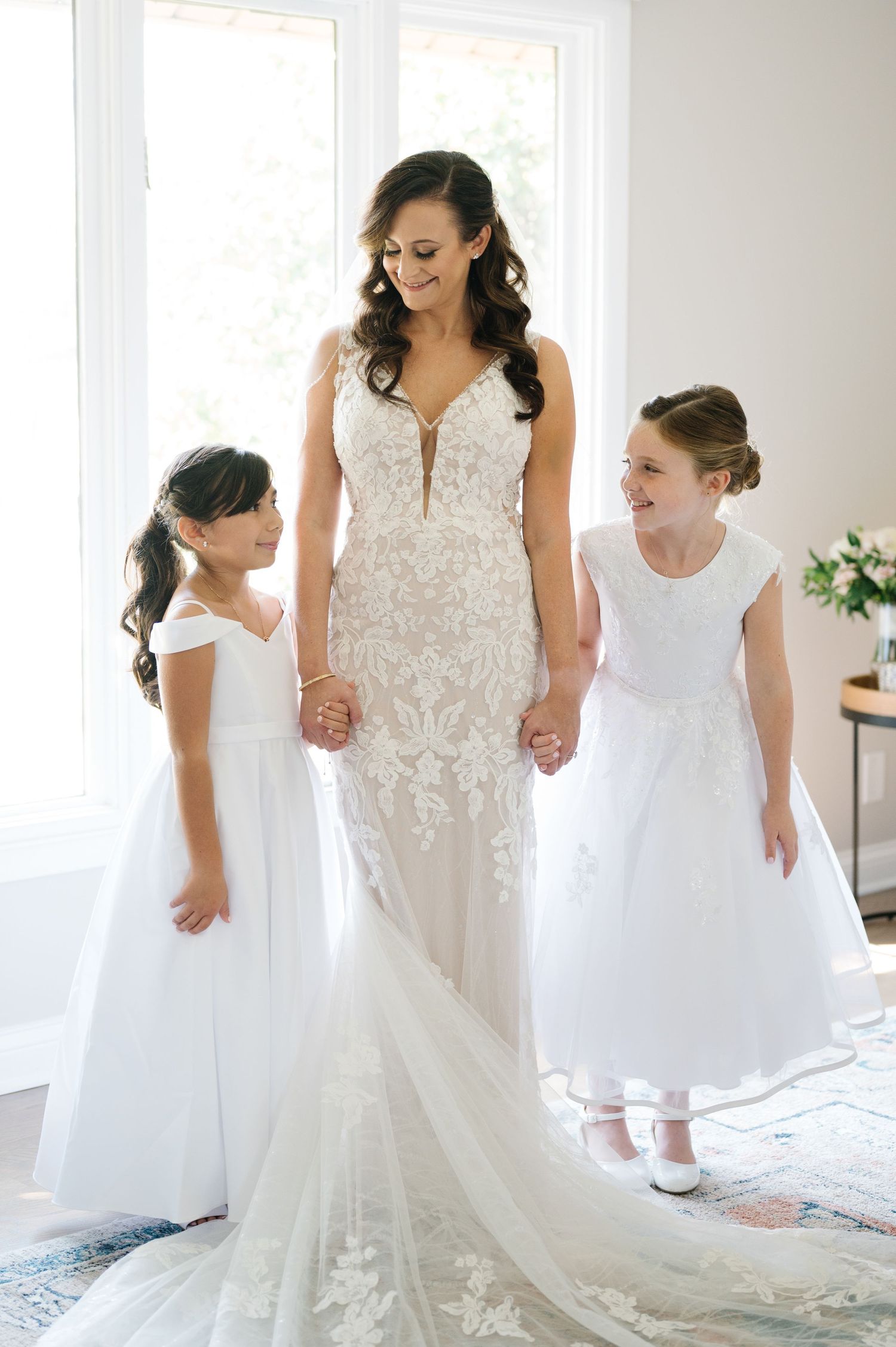 A stunning bridal scene featuring a mermaid-style lace wedding gown with two flower girls in white dresses in a bright room.