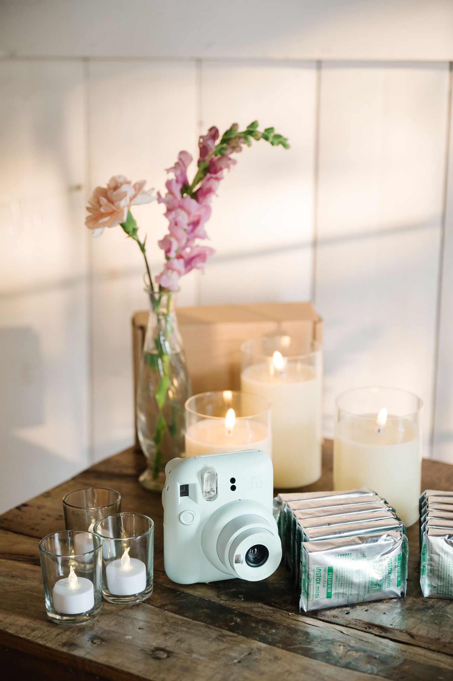 Pastel blue Instax camera beside lit candles and pink flowers in a bottle on rustic wooden table with soft natural lighting.