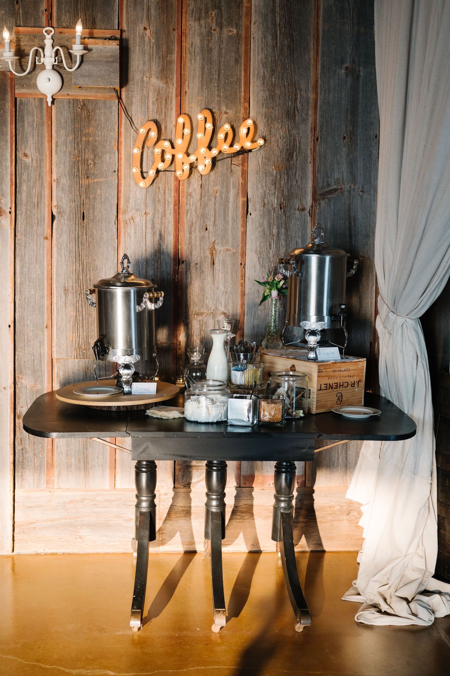 A rustic coffee bar setup with vintage wood walls, a neon coffee sign, and silver coffee urns on an antique black table.