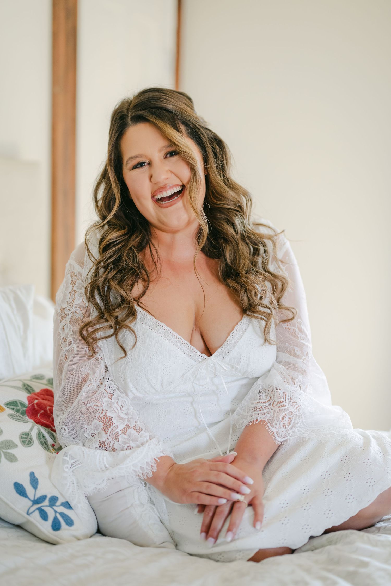 Smiling bride in white lace robe sitting on bed with curly hair, radiating joy on her wedding day. - Youngblood Vineyard Wedding