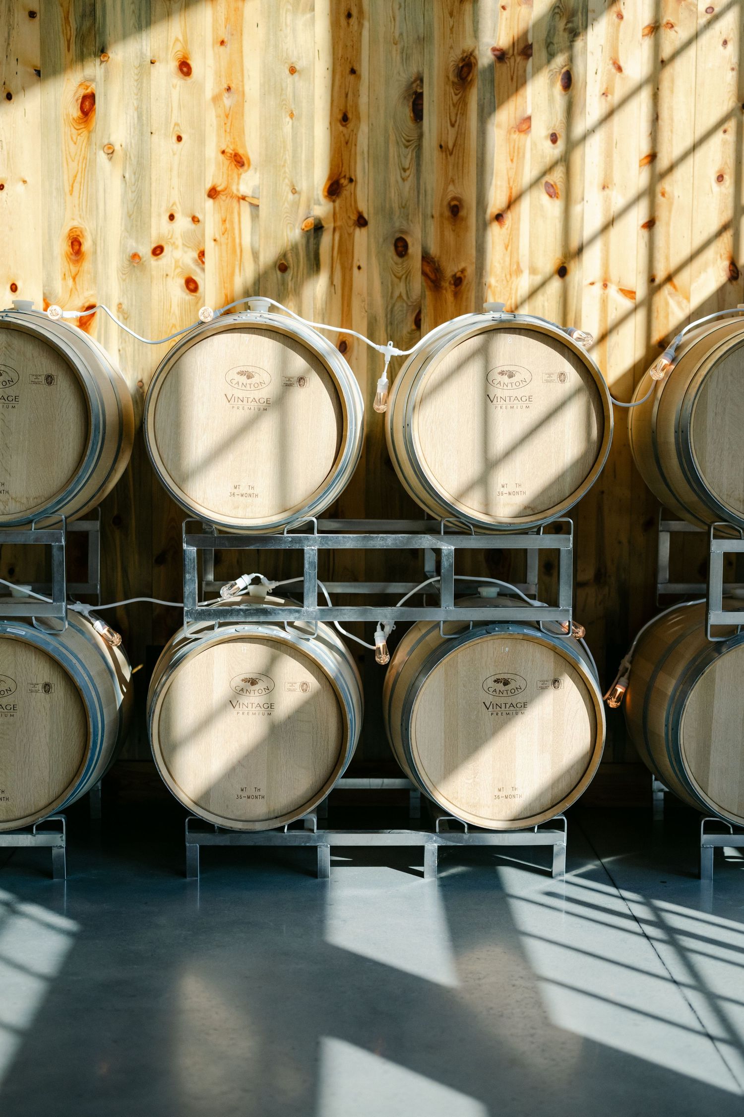 Oak wine barrels stacked on metal racks against a wooden wall in a sunlit winery cellar. - Youngblood Vineyard Wedding