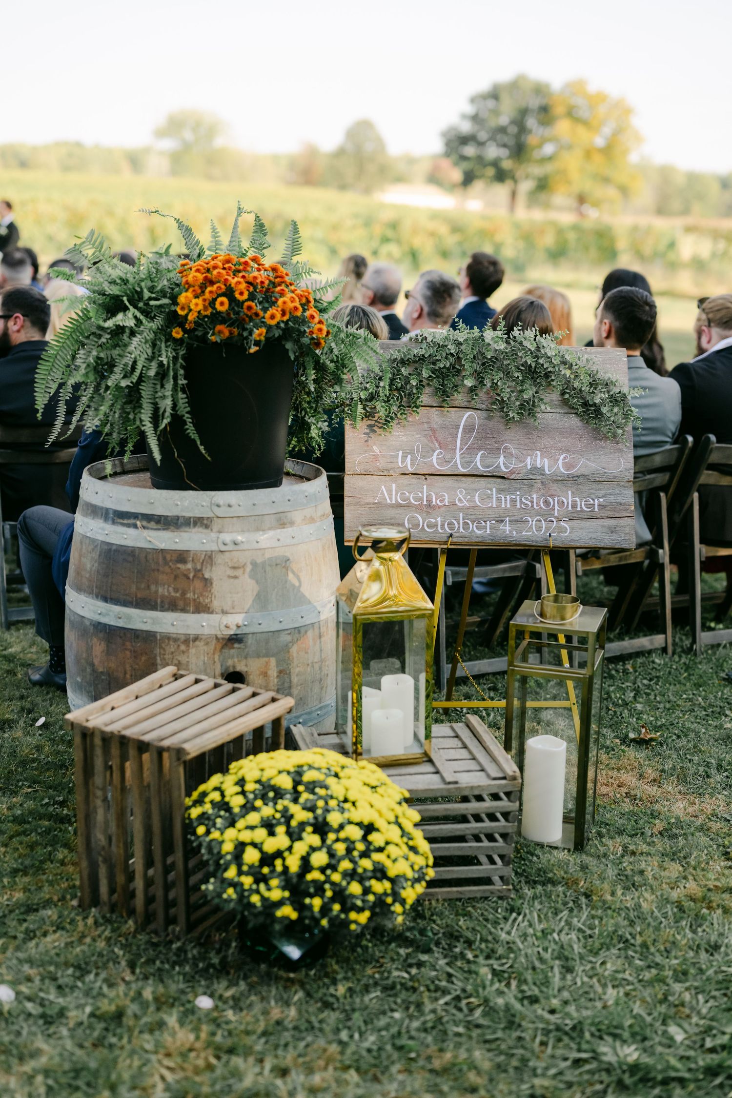 Rustic outdoor wedding ceremony with barrel, welcome sign, lanterns, ferns, and yellow and orange mums. - Youngblood Vineyard Wedding