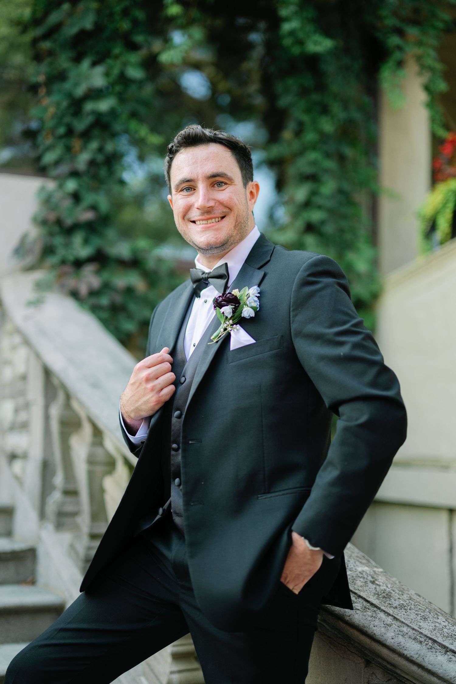 A groom in a classic black tuxedo poses outdoors against a backdrop of lush green foliage. - Meyer's Castle Events