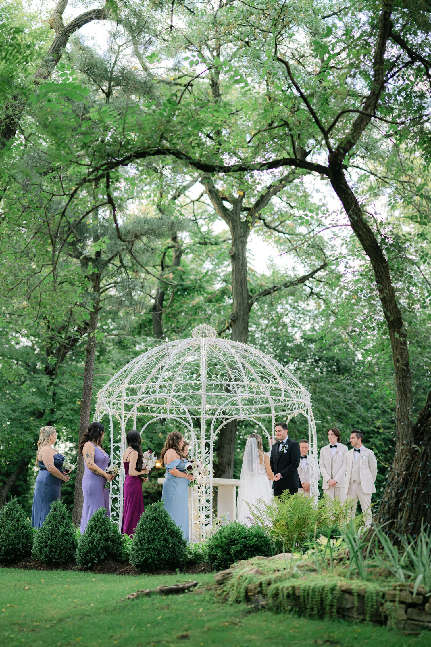 Outdoor wedding ceremony under white gazebo surrounded by lush green trees and guests in colorful attire. - Meyer's Castle Events