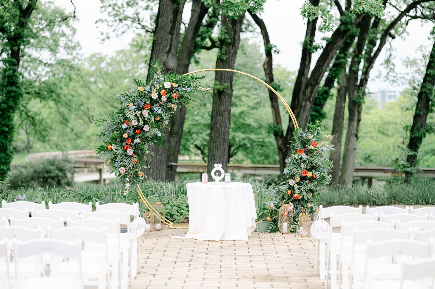 White wooden chairs arranged in rows for an outdoor wedding ceremony under tall trees with orange floral decor. - Hyatt Lodge Oak Brook