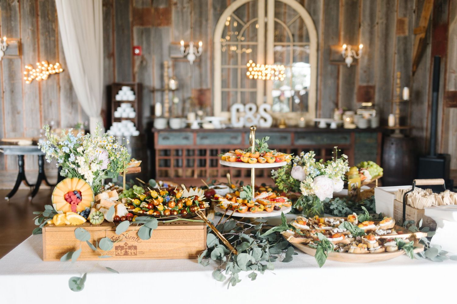 Elegant wedding reception dessert table with tiered serving platters, fresh flowers and greenery in a rustic venue.