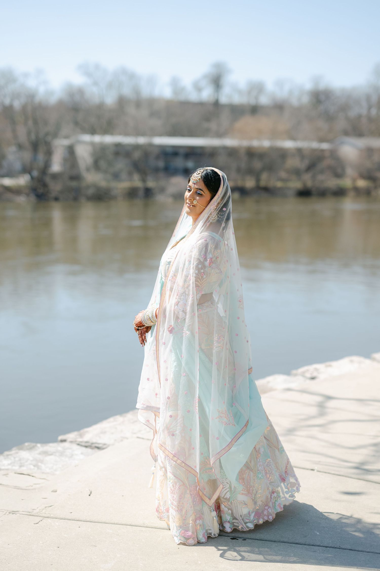 A figure in a flowing white lace dress stands gracefully by a serene lake on a sunny winter day. - The Brix on the Fox