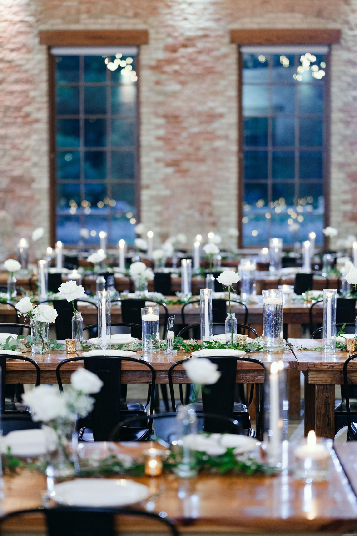 Elegant wedding reception tables with white floral centerpieces and candles in a rustic brick-walled venue.