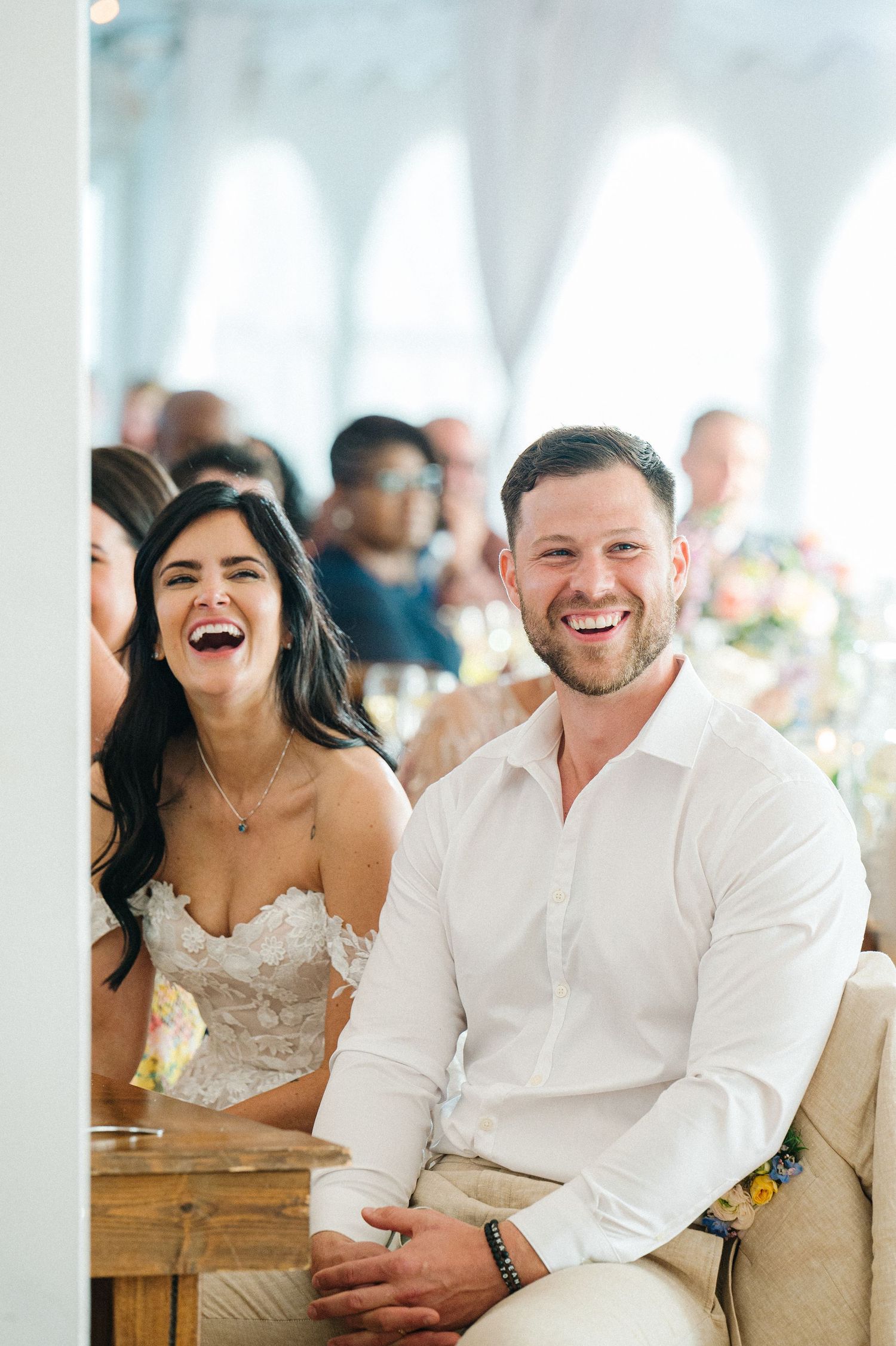 A joyous couple smiles and laughs together at an elegant reception with white arches and warm lighting in the background.