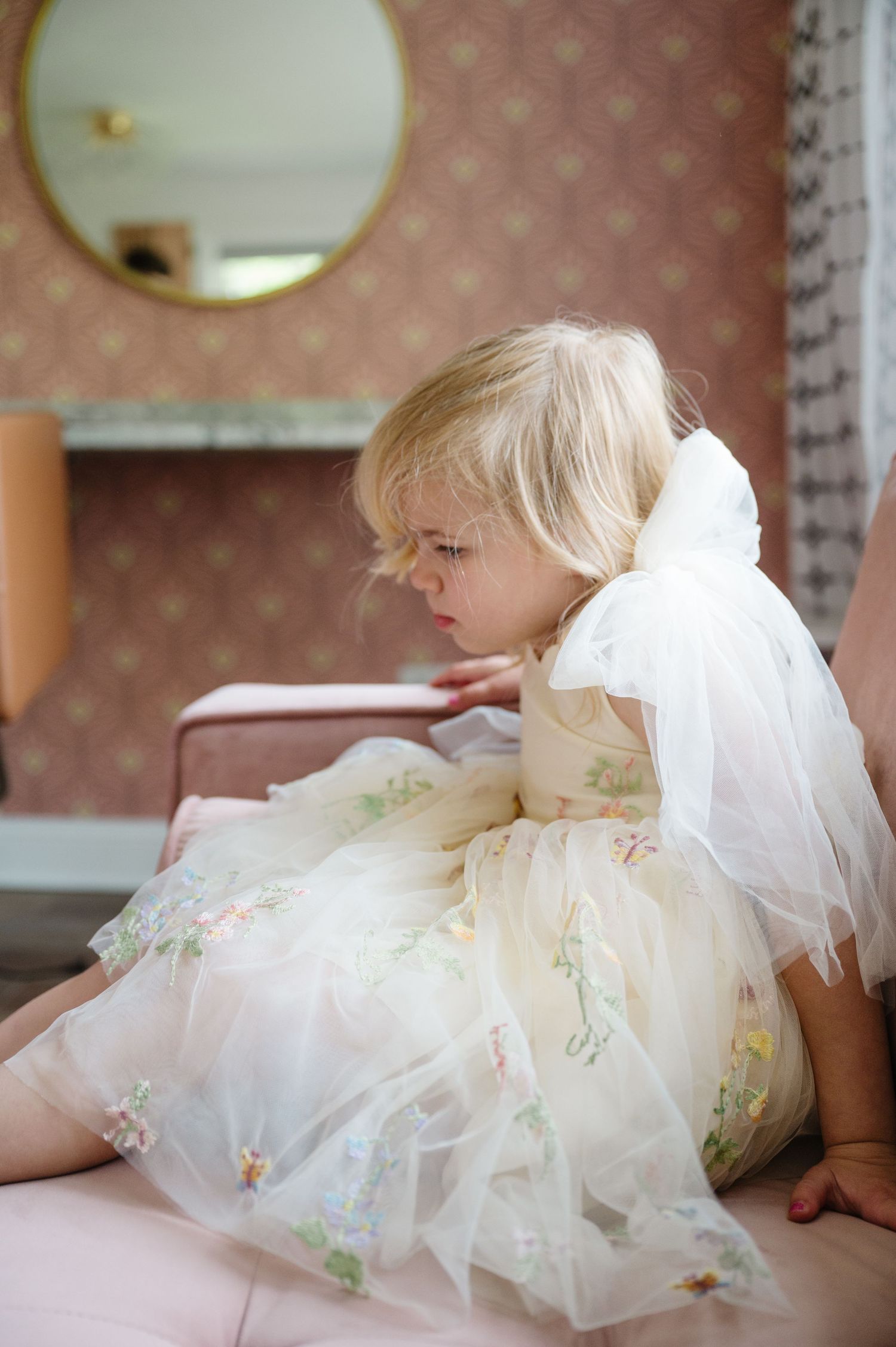 A young child wearing a delicate floral tulle dress and veil sits thoughtfully on a pink couch.