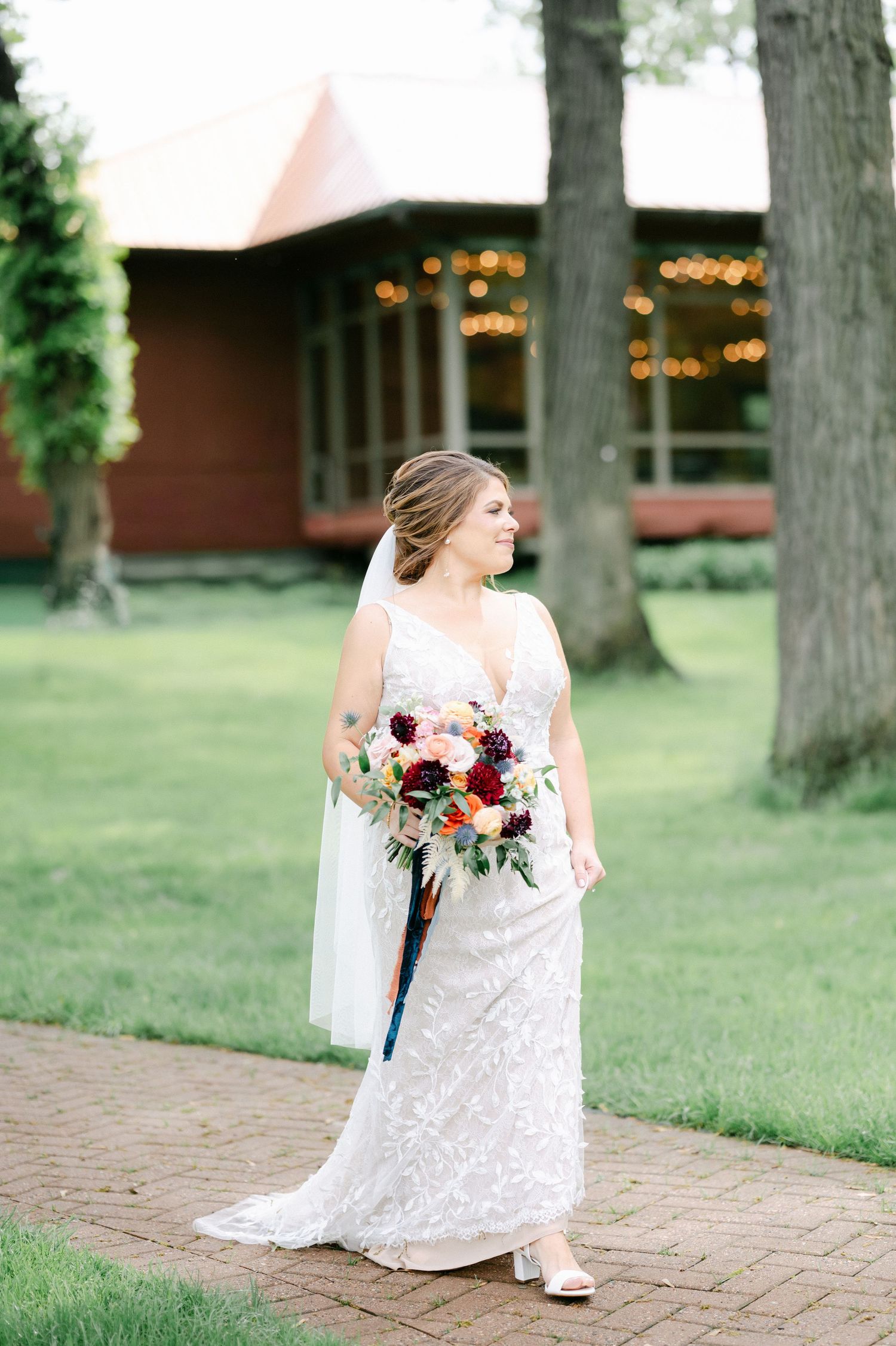 A bride in a lace wedding dress walks on a brick path holding a burgundy and blush floral bouquet. - Hyatt Lodge Oak Brook