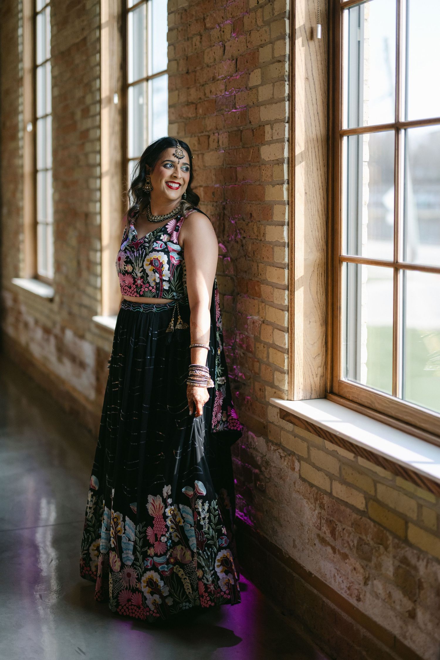 A person in a floral black dress poses by large windows in a rustic brick-walled room with natural lighting. - The Brix on the Fox